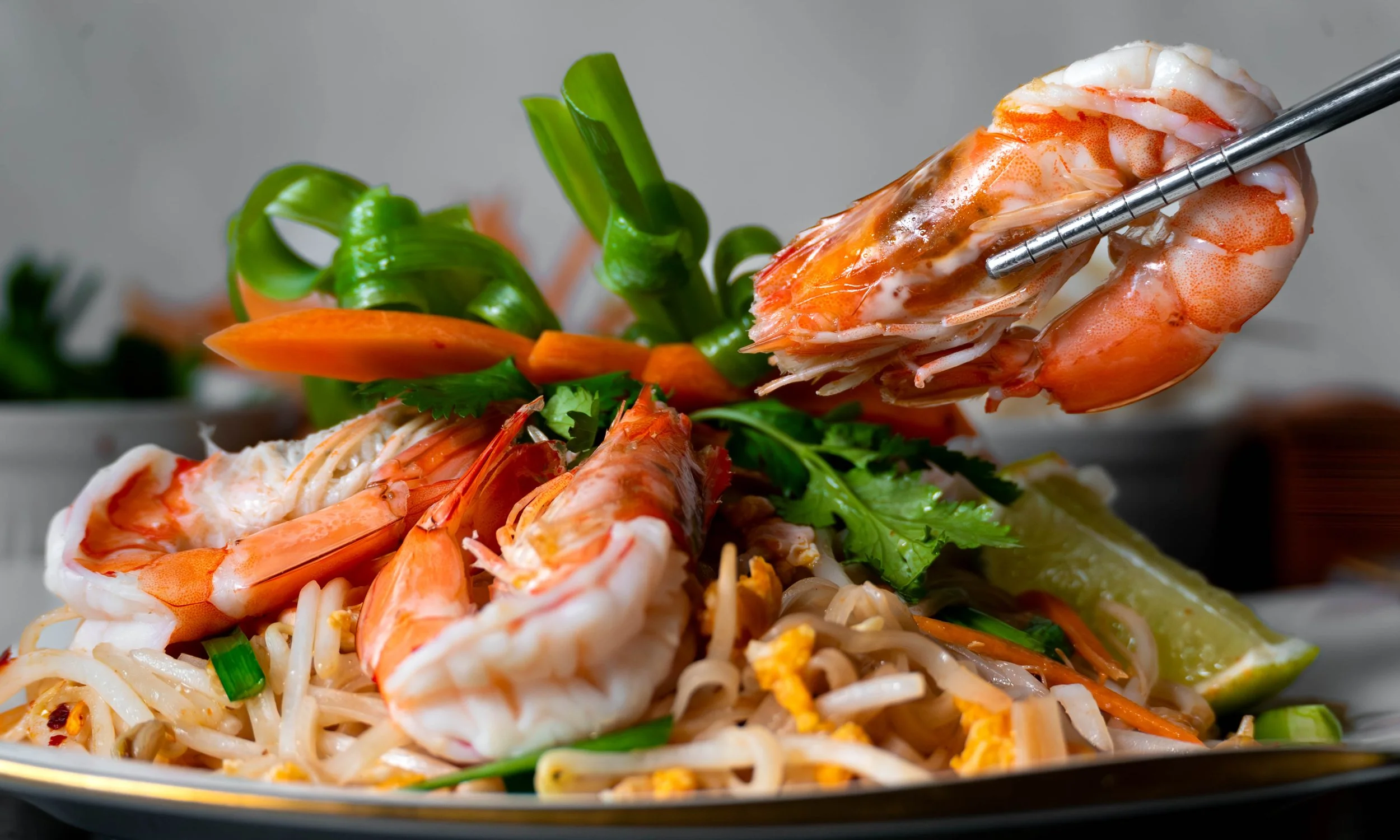Close-up of a plate of shrimp pad thai with vegetables and lime wedge, with chopsticks picking up a shrimp.