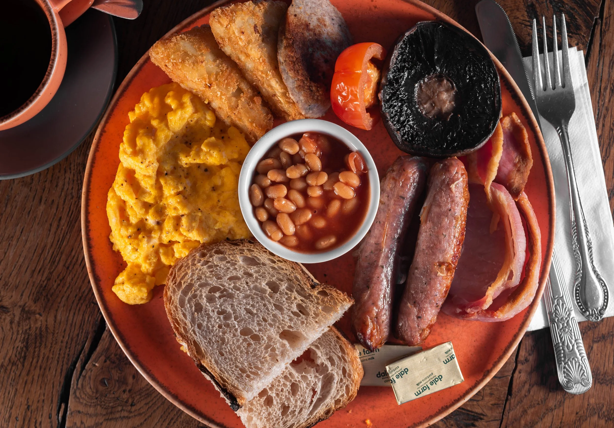 A traditional English breakfast on a round orange plate, including scrambled eggs, baked beans, grilled tomatoes, black pudding, sausages, bacon, toasted bread, and fried potato wedges, next to a cup of black coffee.