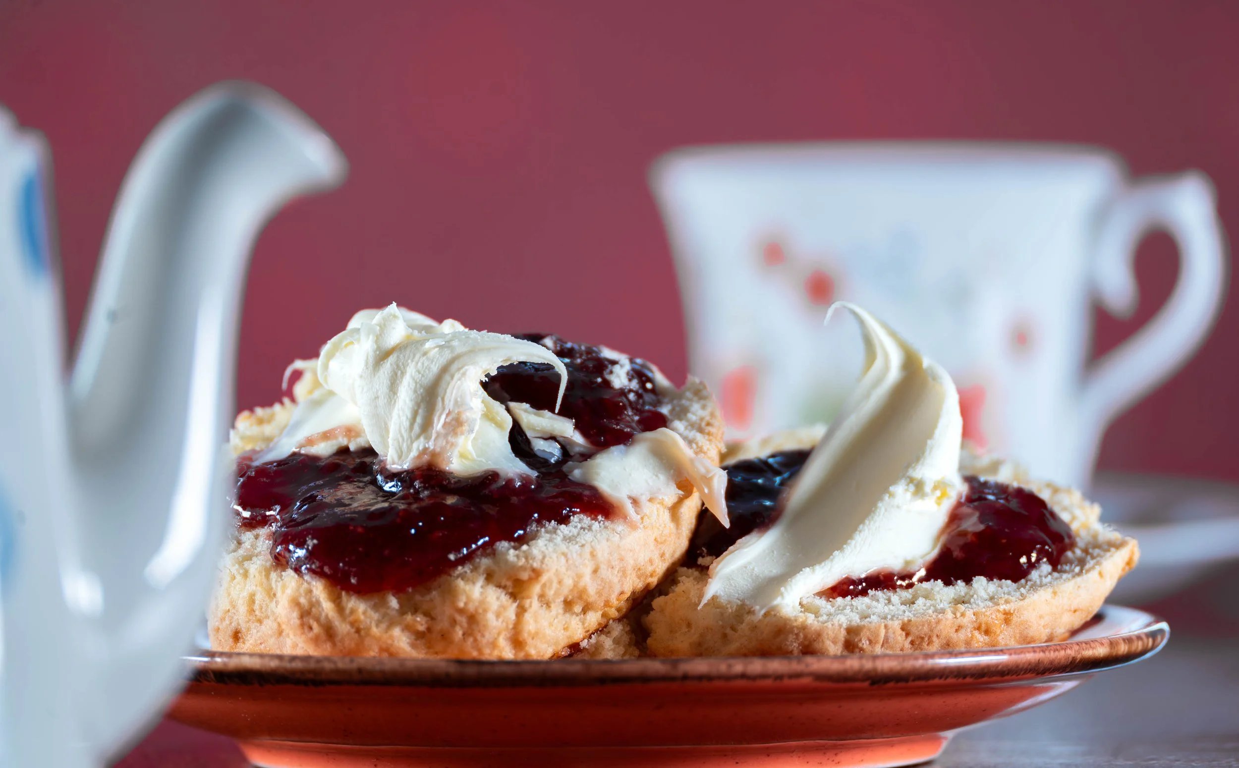 Close-up of two scones with cream and jam on a plate, with a teapot and teacup in the background.