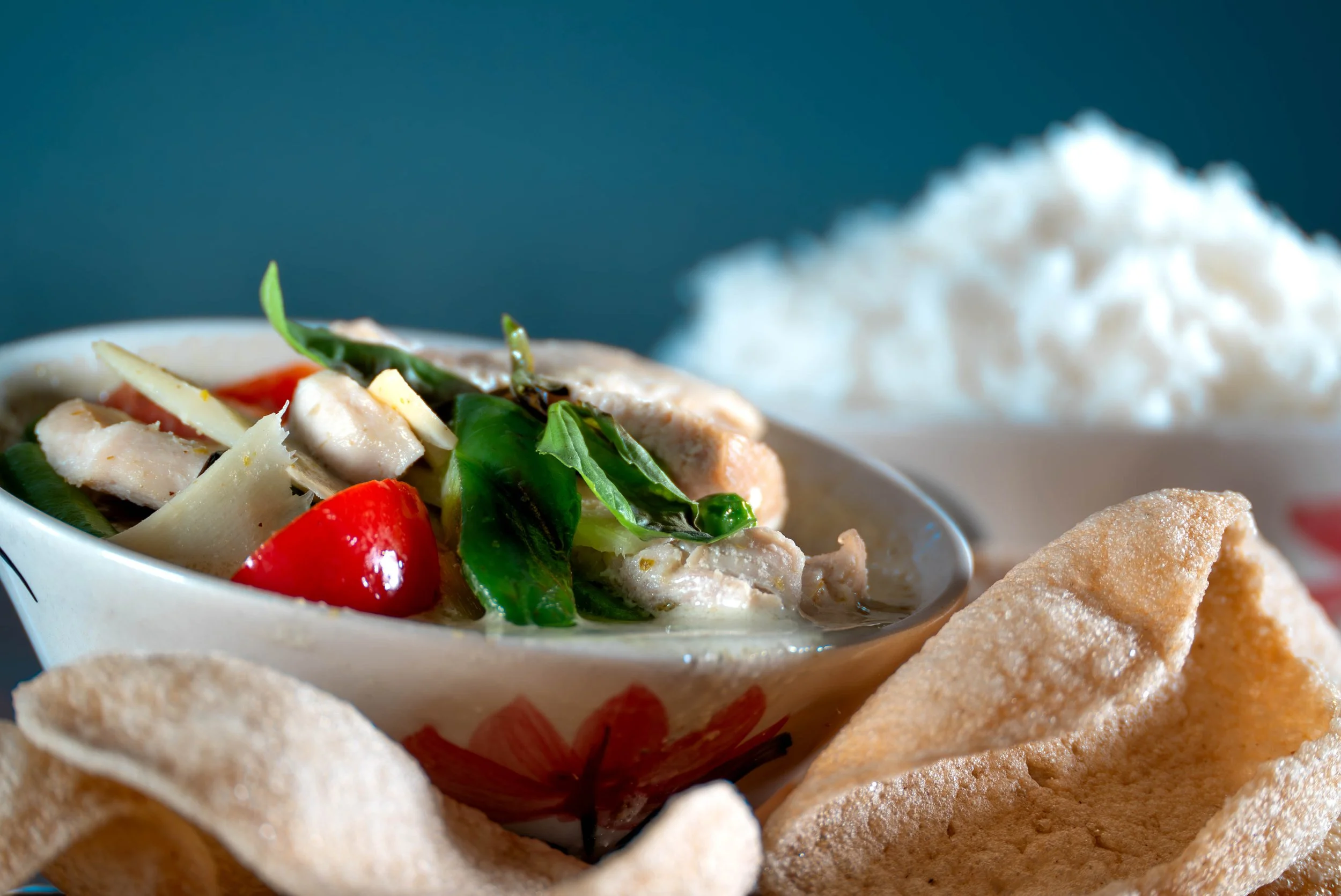 A bowl of chicken salad with fresh vegetables, surrounded by prawn crackers and a bowl of rice in the background.
