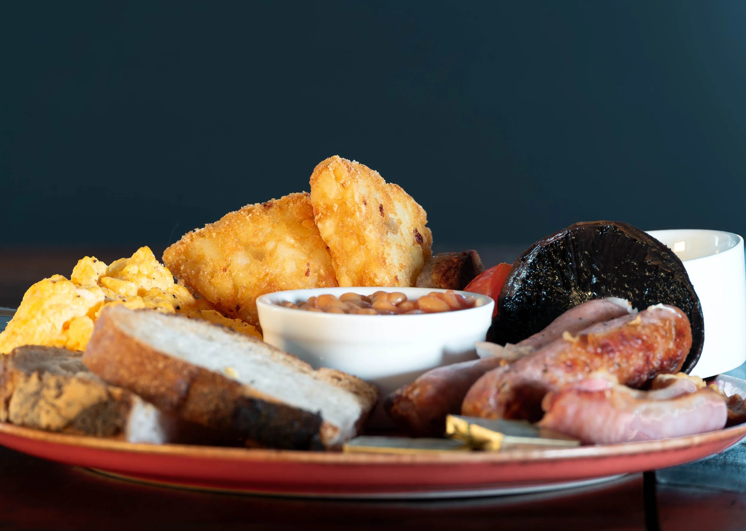 Full English breakfast on a pottery plate, including scrambled eggs, fried bread, baked beans, hash browns, grilled tomatoes, baked mushrooms, rashers of bacon, and a cup of tea or coffee.