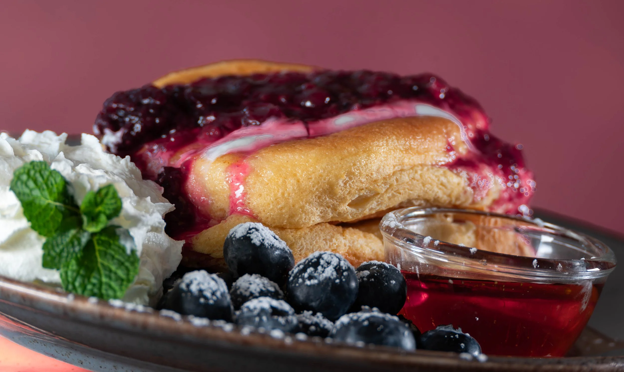 Close-up of a blueberry pancake stack topped with berry compote, served with whipped cream, blueberries, and a small bowl of syrup on a plate.