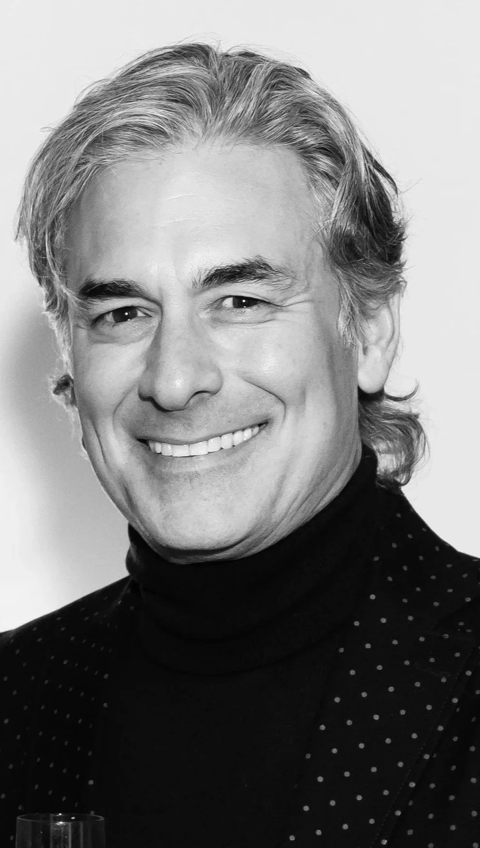 A smiling middle-aged man with wavy gray hair, wearing a black turtleneck with small white polka dots.
