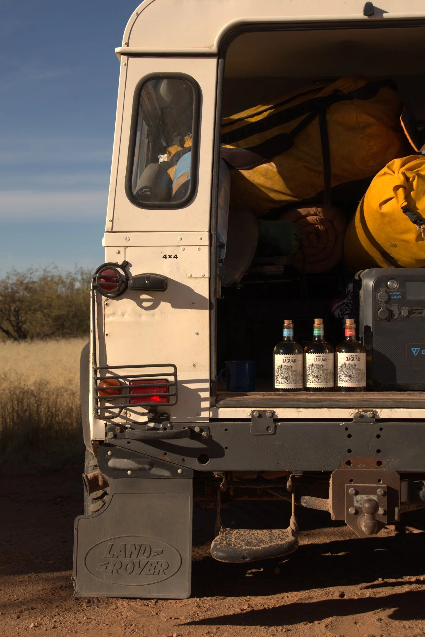 Back of Land Rover vehicle with camping gear, three bottles of Jagua rum, and a small speaker inside the trunk in an outdoor setting.