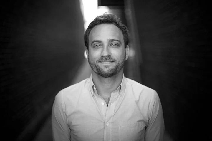 A black and white portrait of a man with short hair and a beard, smiling slightly, wearing a collared shirt, standing outdoors with blurred background.