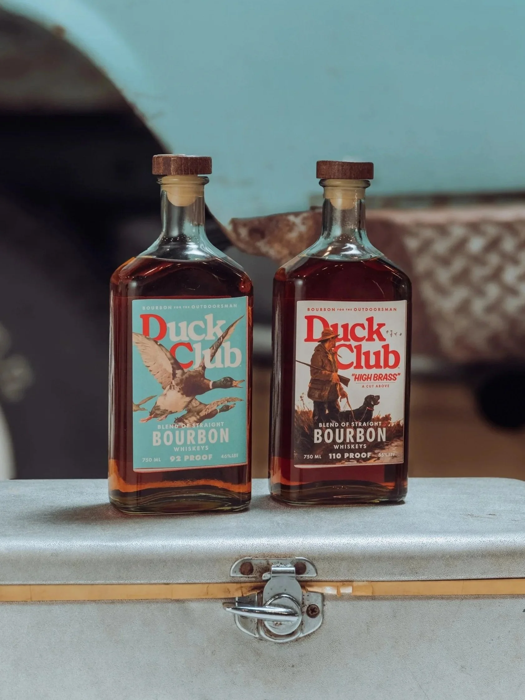 Two bottles of Duck Club bourbon whiskey on a metal surface, one labeled blend of straight bourbon and the other labeled 'High Brass' with an image of a man with a dog, against a blurred background.