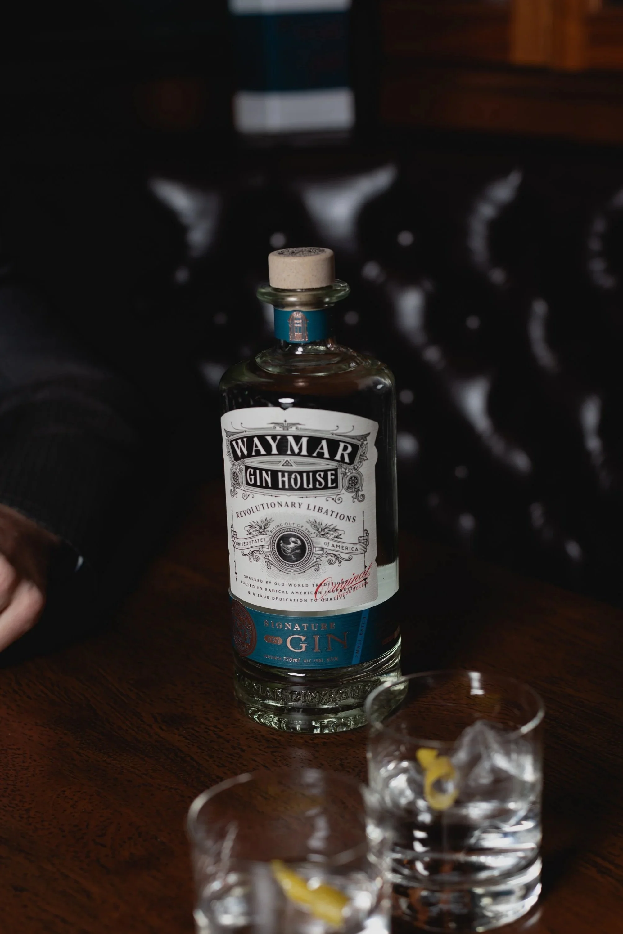 Bottle of Waymar Gin House signature gin on a wooden table with two glasses of ice and lemon wedge, against a dark background.