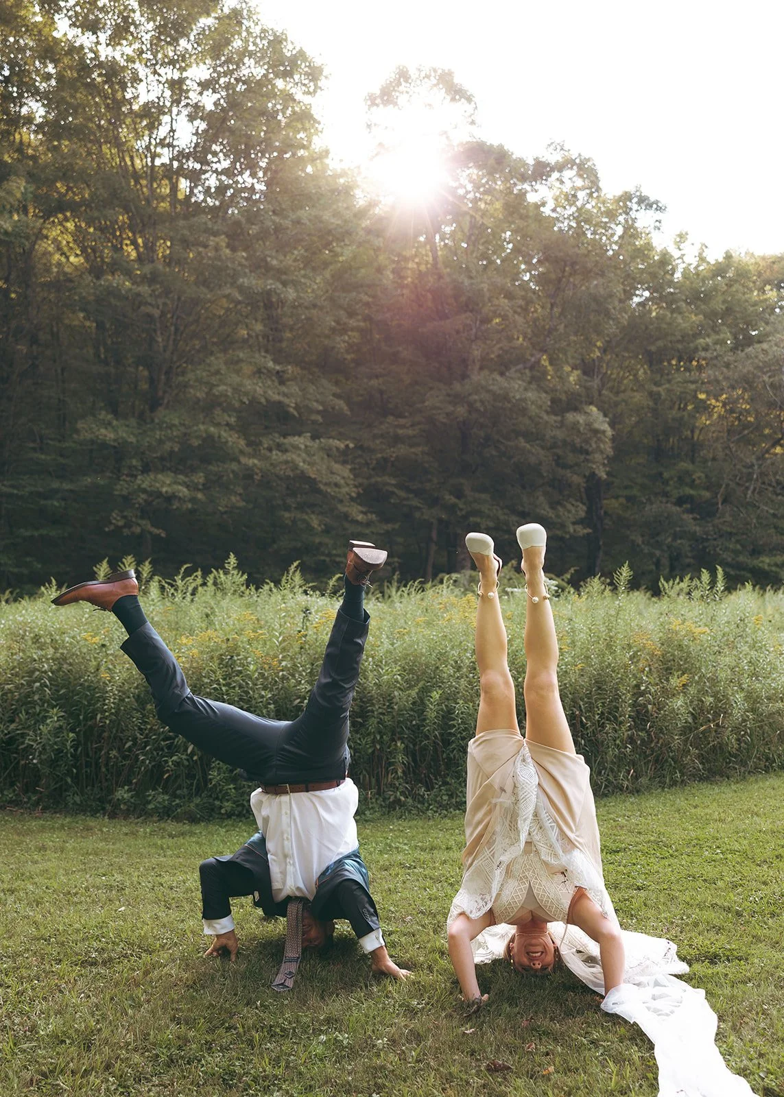 A man and a woman are doing handstands on the grass in a park with tall grass and trees in the background; the man is upside down with his legs in the air, wearing dress shoes and dark pants; the woman is also upside down, wearing a white dress and yellow shoes, and smiling.