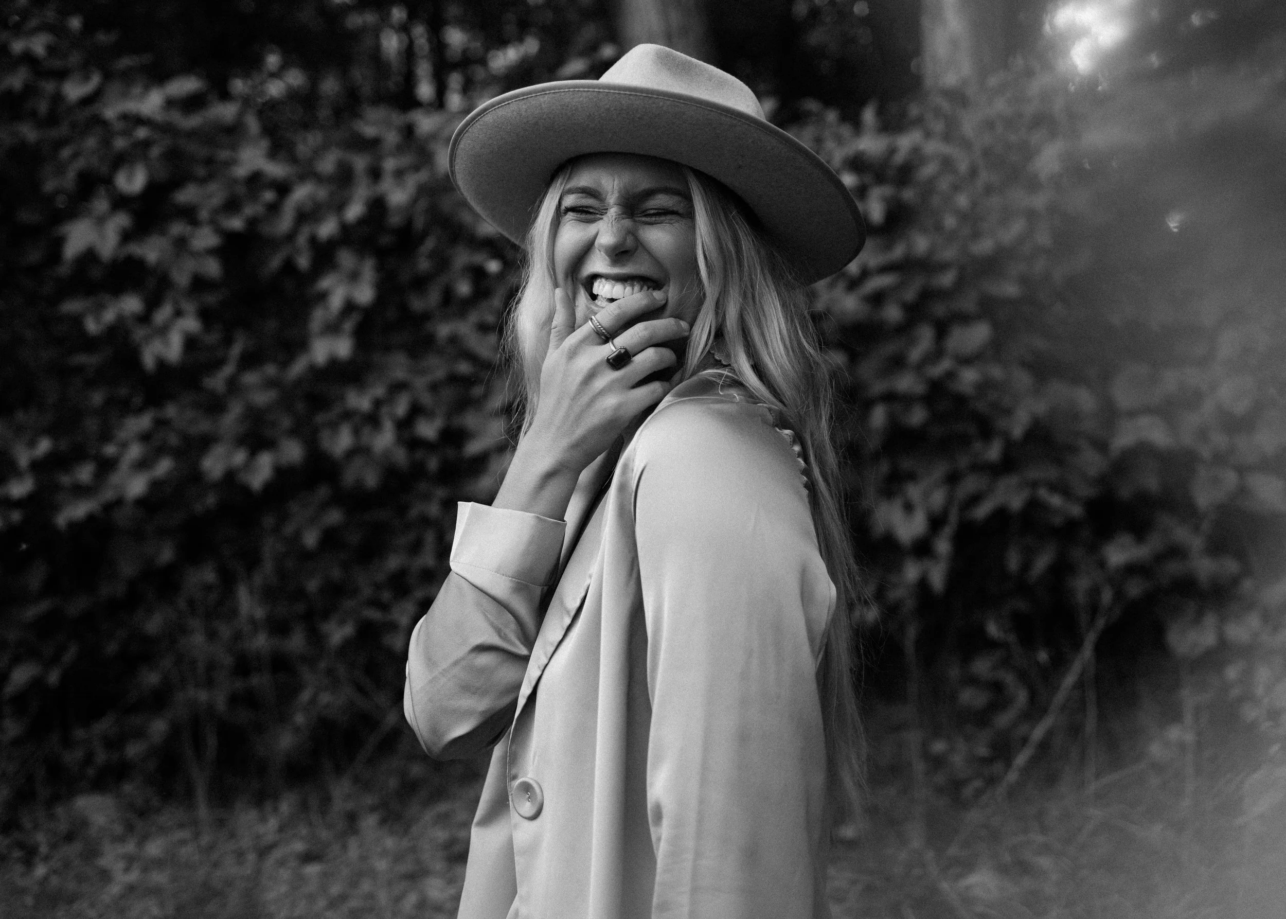 Black and white photo of a woman wearing a wide-brimmed hat, smiling joyfully with her hand near her face, outdoors background with blurred foliage.