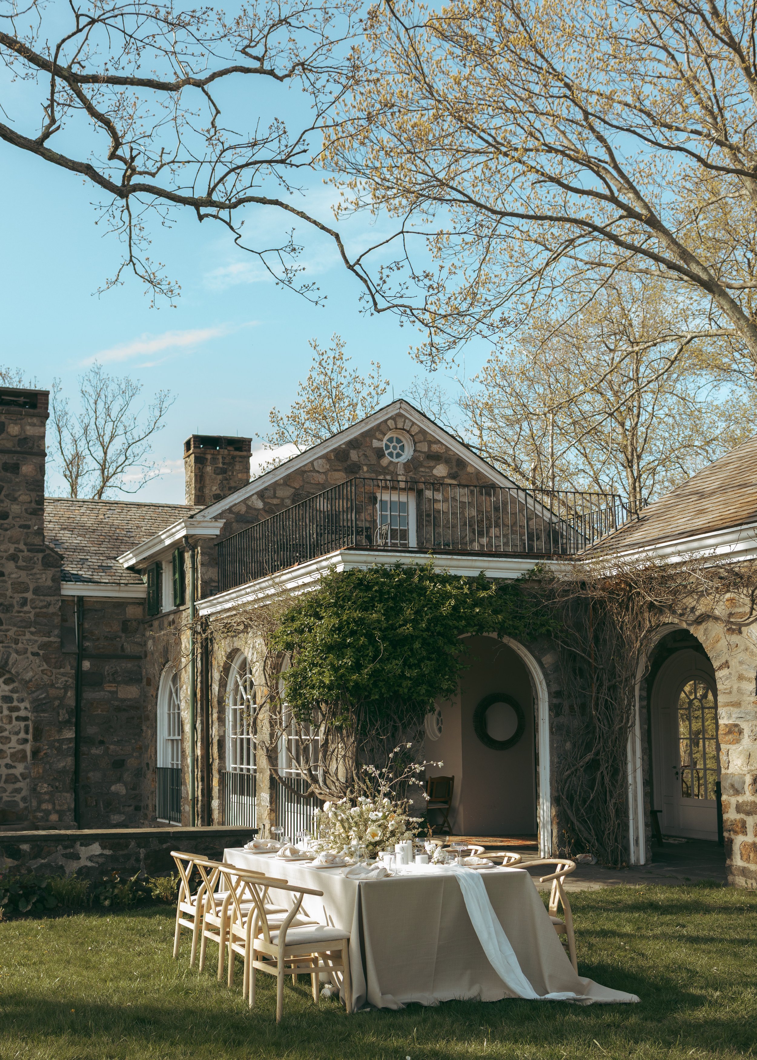 An outdoor gathering setup with a long table covered with a white tablecloth, set with white plates, glasses, and a floral centerpiece. The setting is in a lush grassy yard in front of a stone house with arched windows and a balcony, surrounded by trees with sparse leaves on a sunny day.