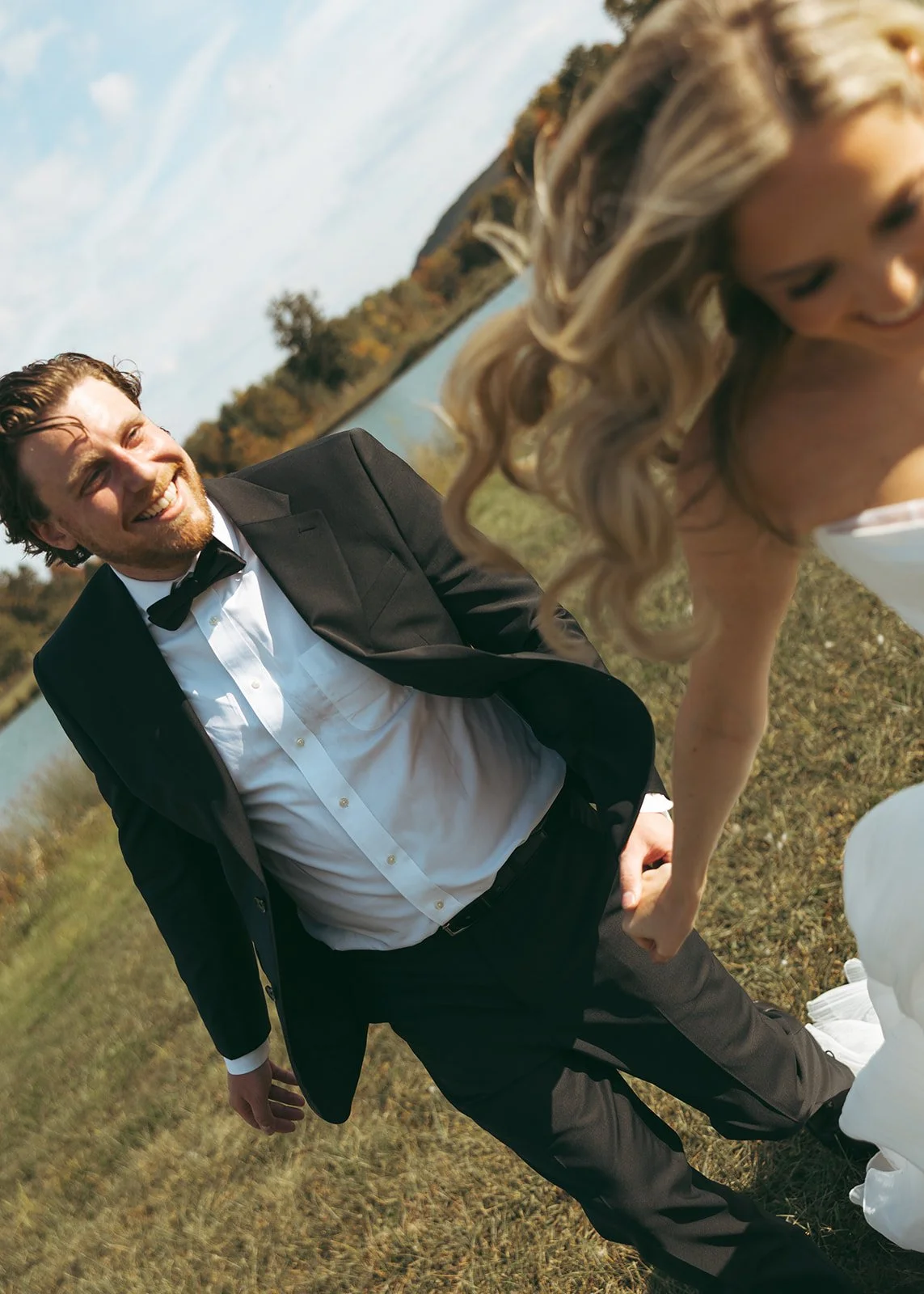 A smiling man in a tuxedo with a bow tie kneeling on the grass next to a blonde woman in a white dress, outdoors near a body of water, with trees in the background.
