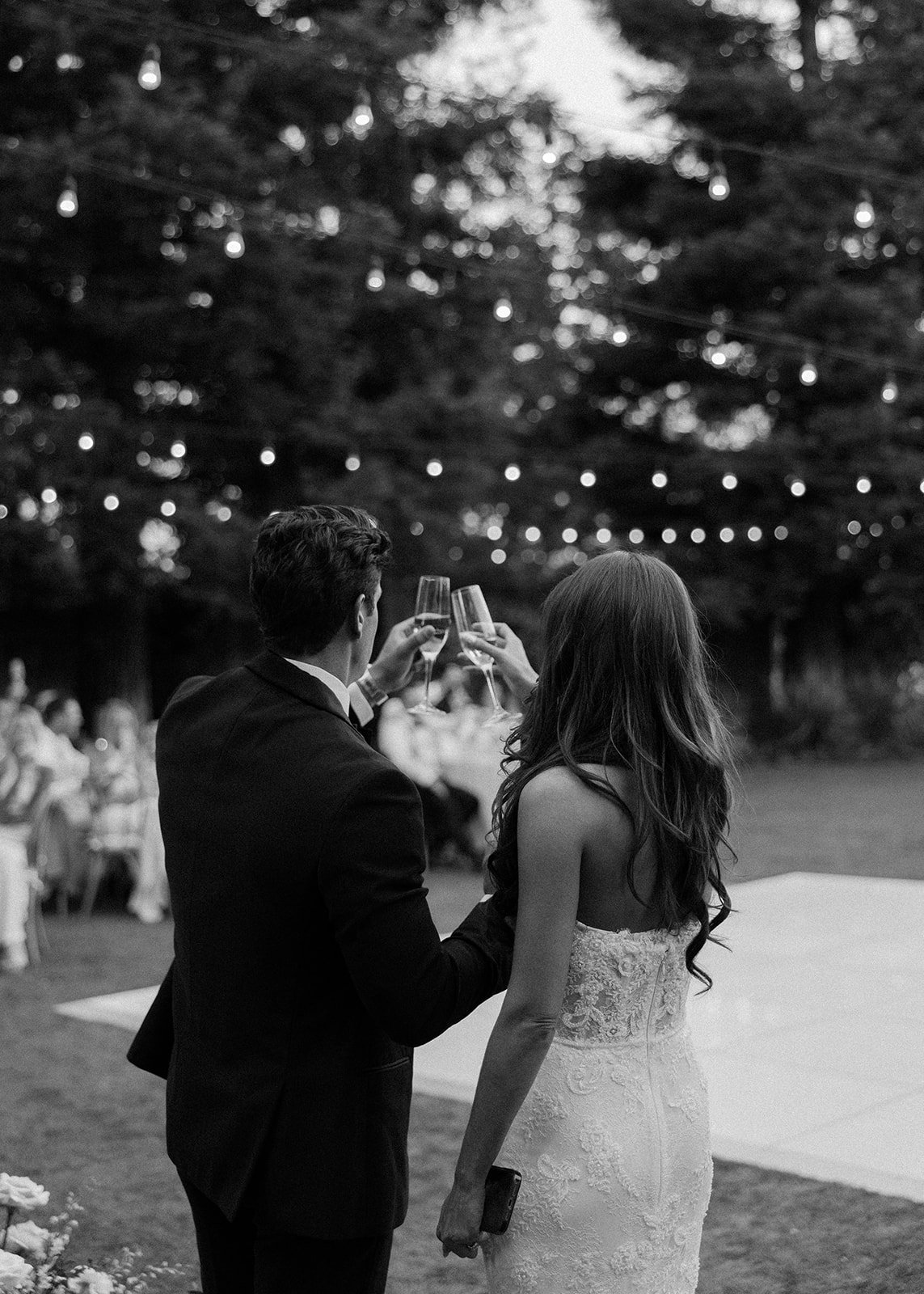 A black and white photo of a man and woman in formal attire holding glasses and toasting at an outdoor wedding reception, with string lights hanging overhead and tables visible in the background.