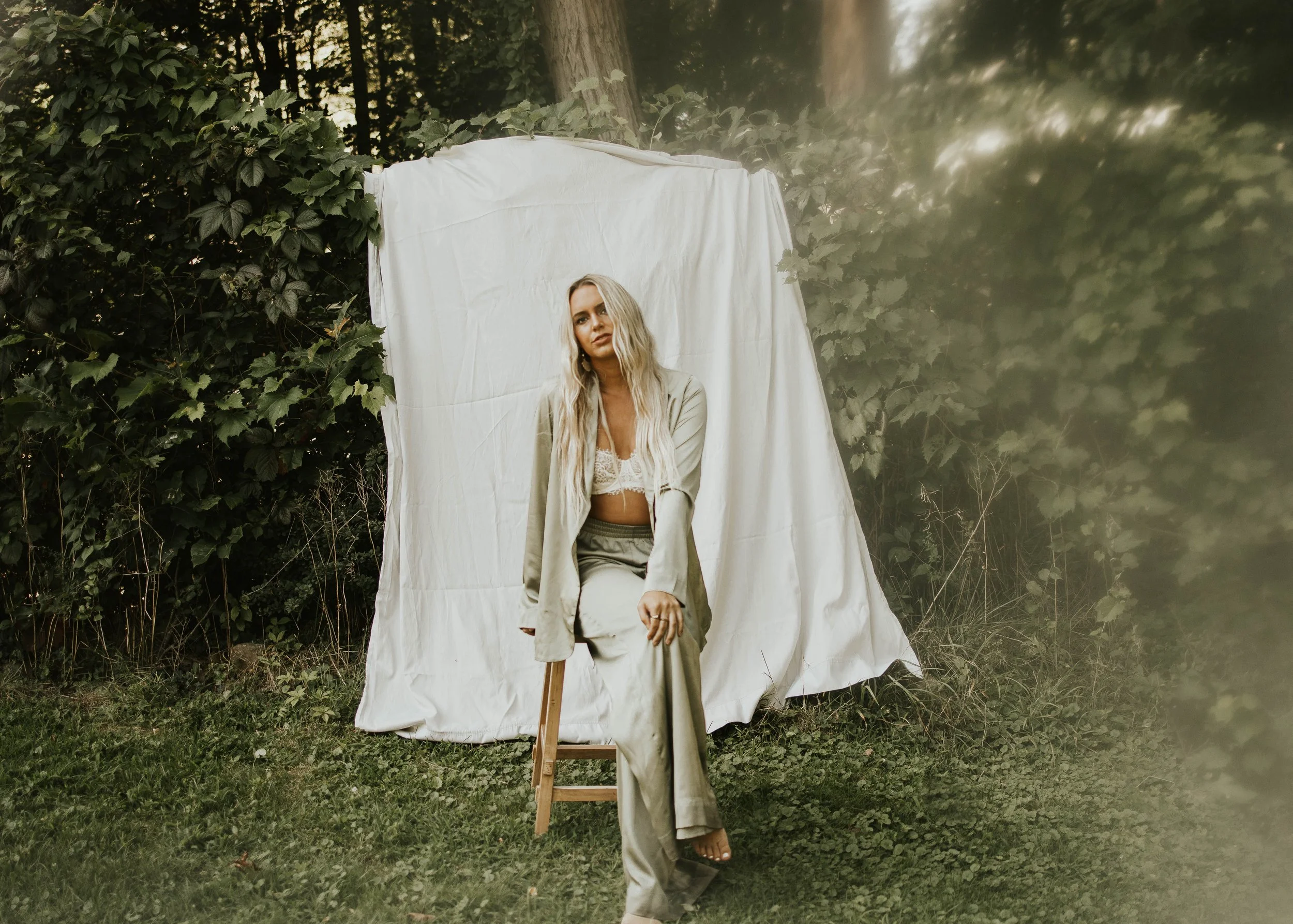 A woman with long blonde hair sitting on a wooden stool outdoors in front of a white cloth backdrop, surrounded by green foliage and trees.
