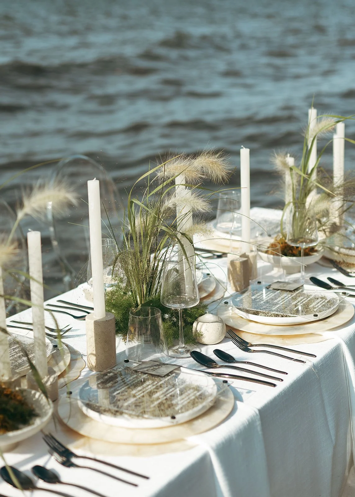 Elegant outdoor table setup with white tablecloth, greenery, and candles on a lakeside.