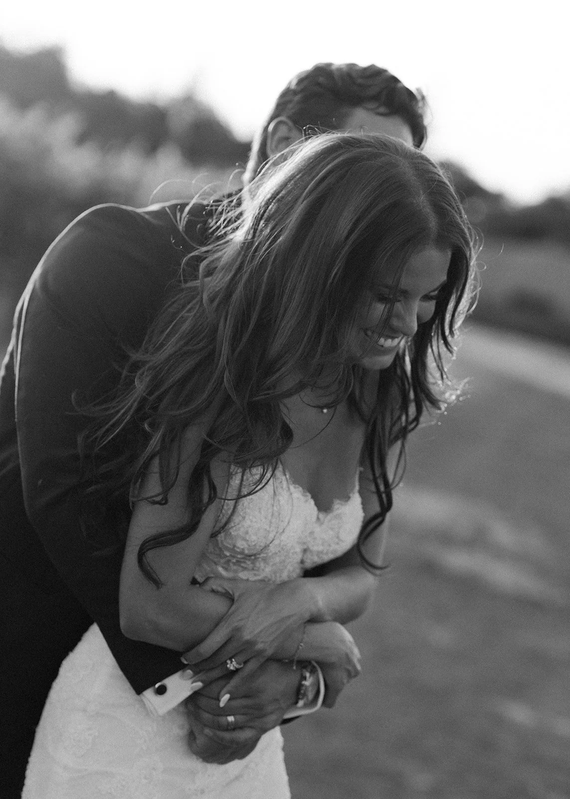 Bride smiling as groom embraces her from behind during golden hour, capturing an intimate and romantic wedding portrait.