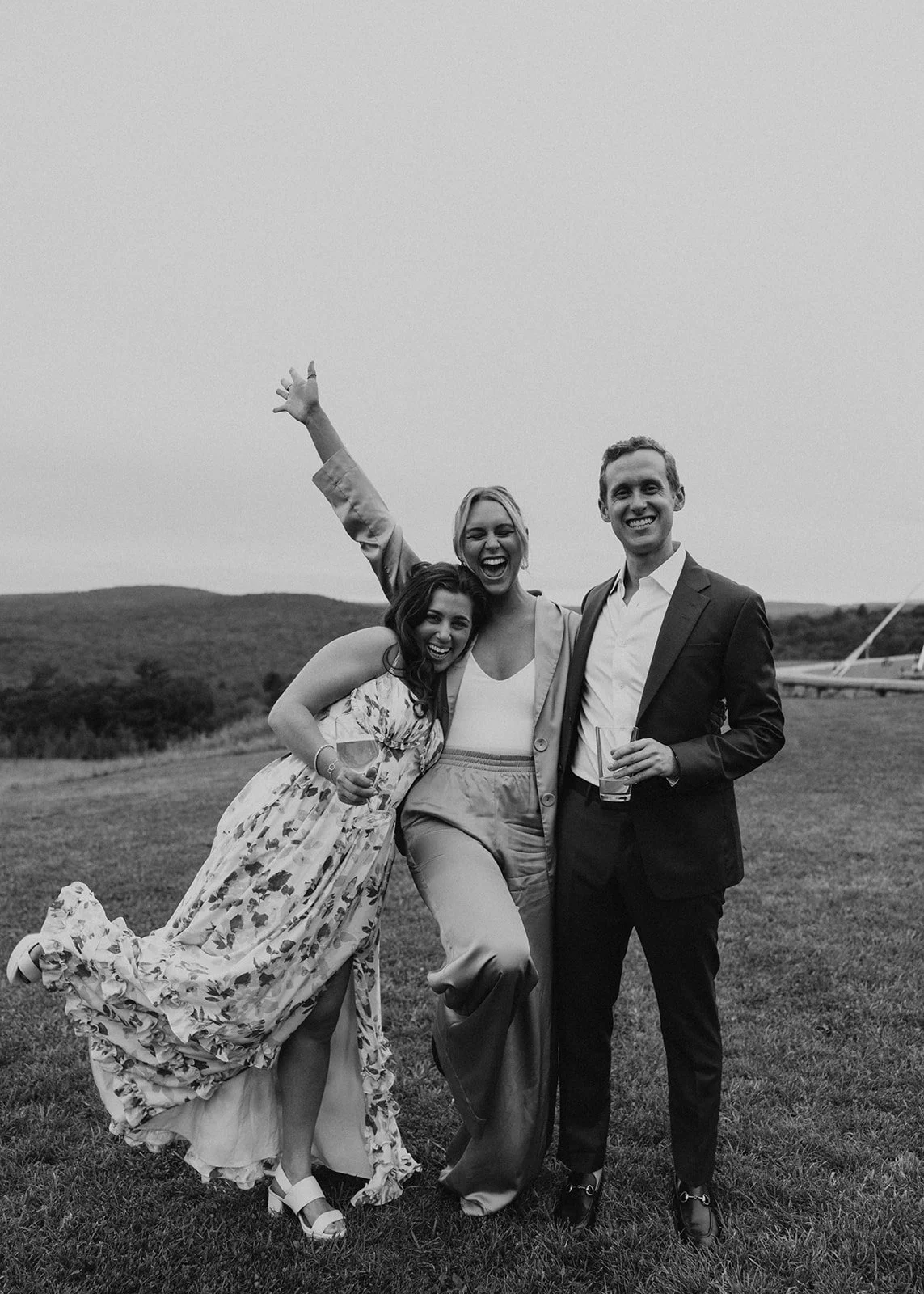 Three wedding guests celebrating together on a scenic hillside, laughing and holding drinks during a wedding reception.