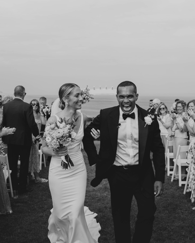 A newlywed couple walks down the aisle smiling, with guests clapping and celebrating outdoors near the coast.