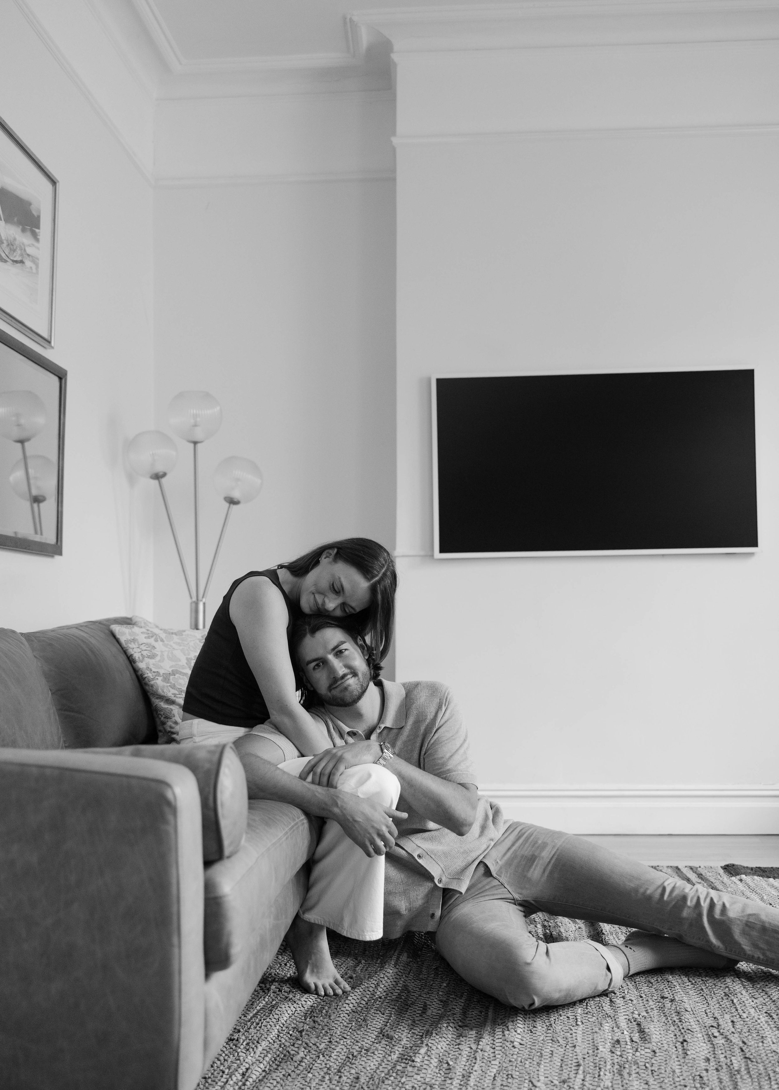 A black and white photo of a woman leaning over a man sitting on the floor next to a sofa in a living room. The woman smiles while resting her head on his shoulder, and the man looks at the camera with a slight smile. There are framed pictures on the wall, a floor lamp with multiple bulbs, and a large TV mounted on the wall.