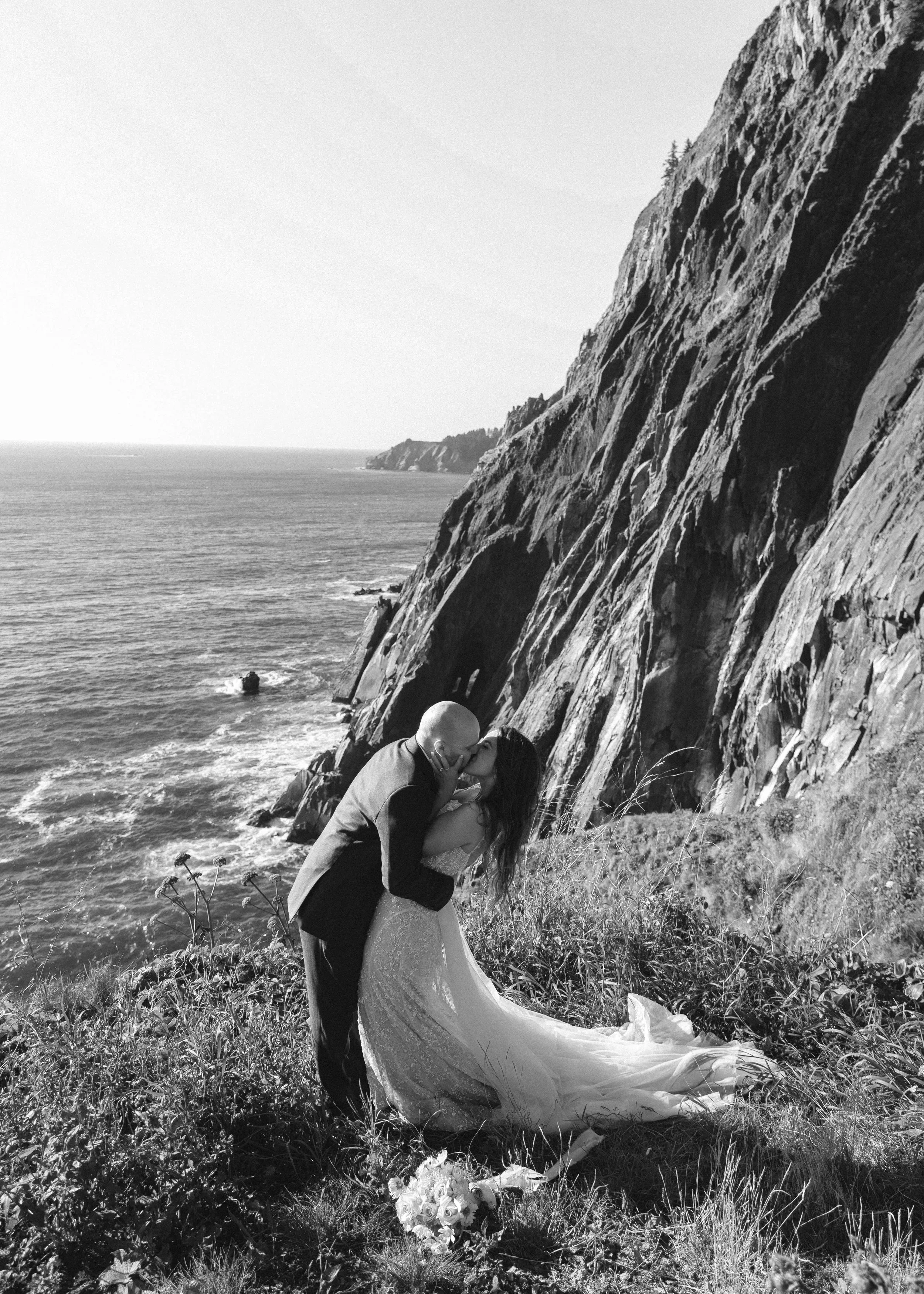 A black and white photo of a couple kissing on a grassy cliffside near the ocean, with rocky cliffs in the background and a bouquet of flowers on the ground.