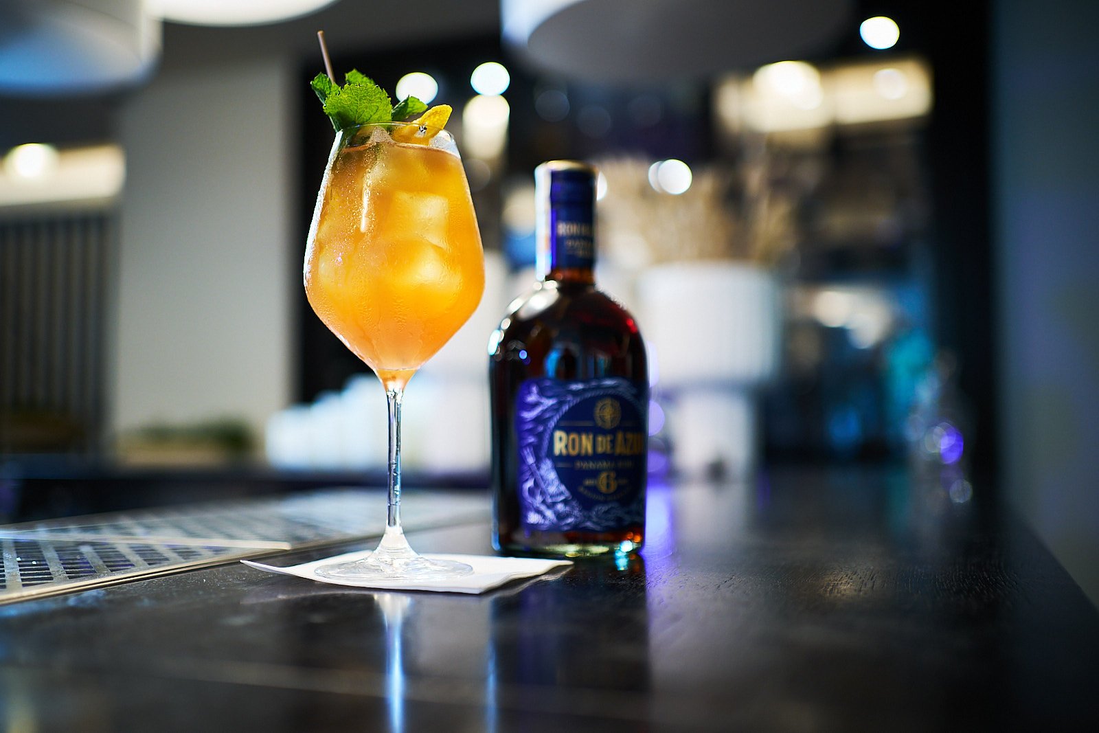An orange cocktail garnished with mint and lemon slices, placed on a bar counter next to a bottle of Ron de AZÚL rum.