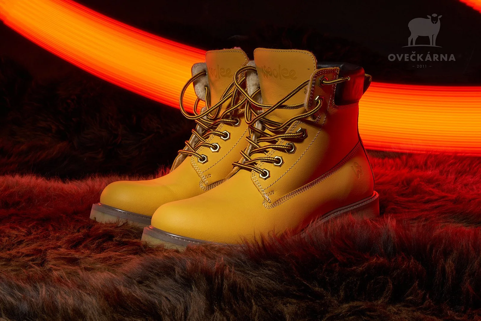 A pair of yellow leather hiking boots with black and yellow laces, set on a faux fur surface with a red and orange neon light in the background.