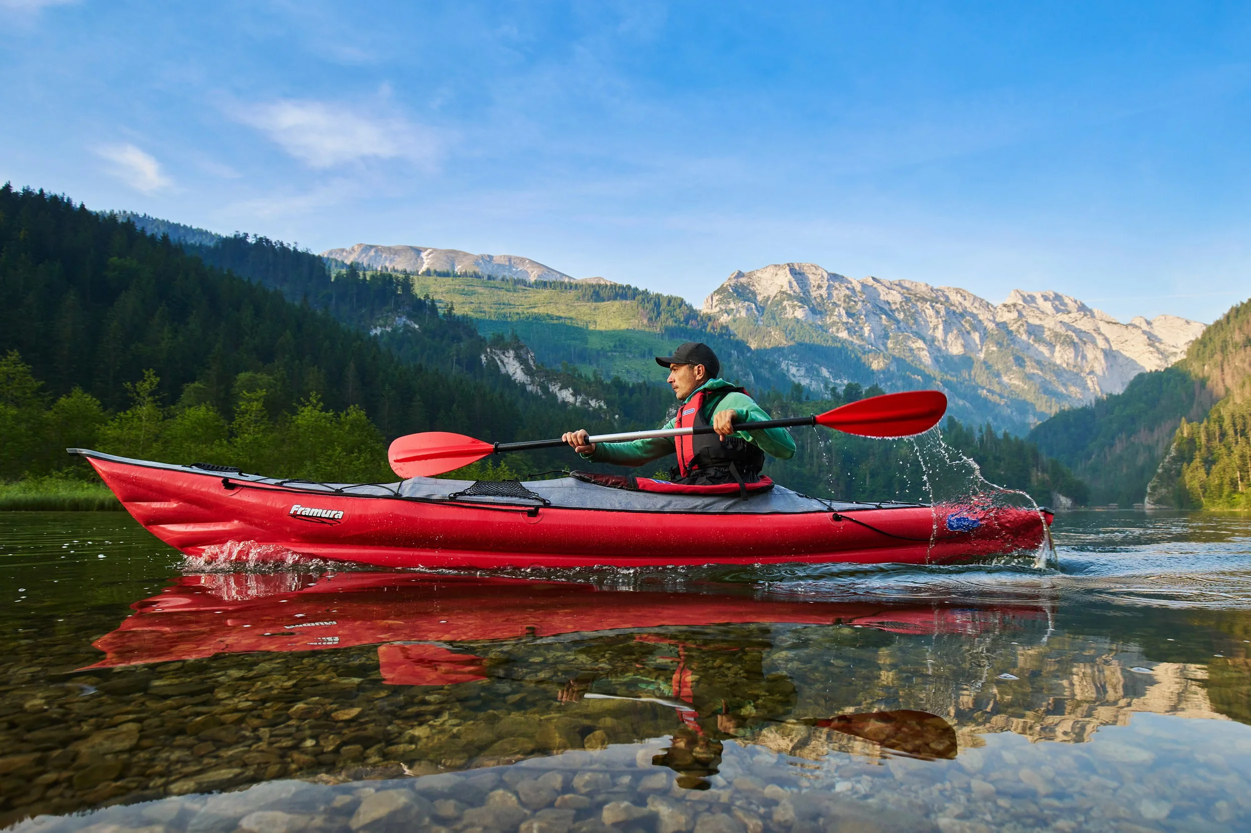 A person kayaking on a calm river with a scenic mountainous landscape and clear blue sky.