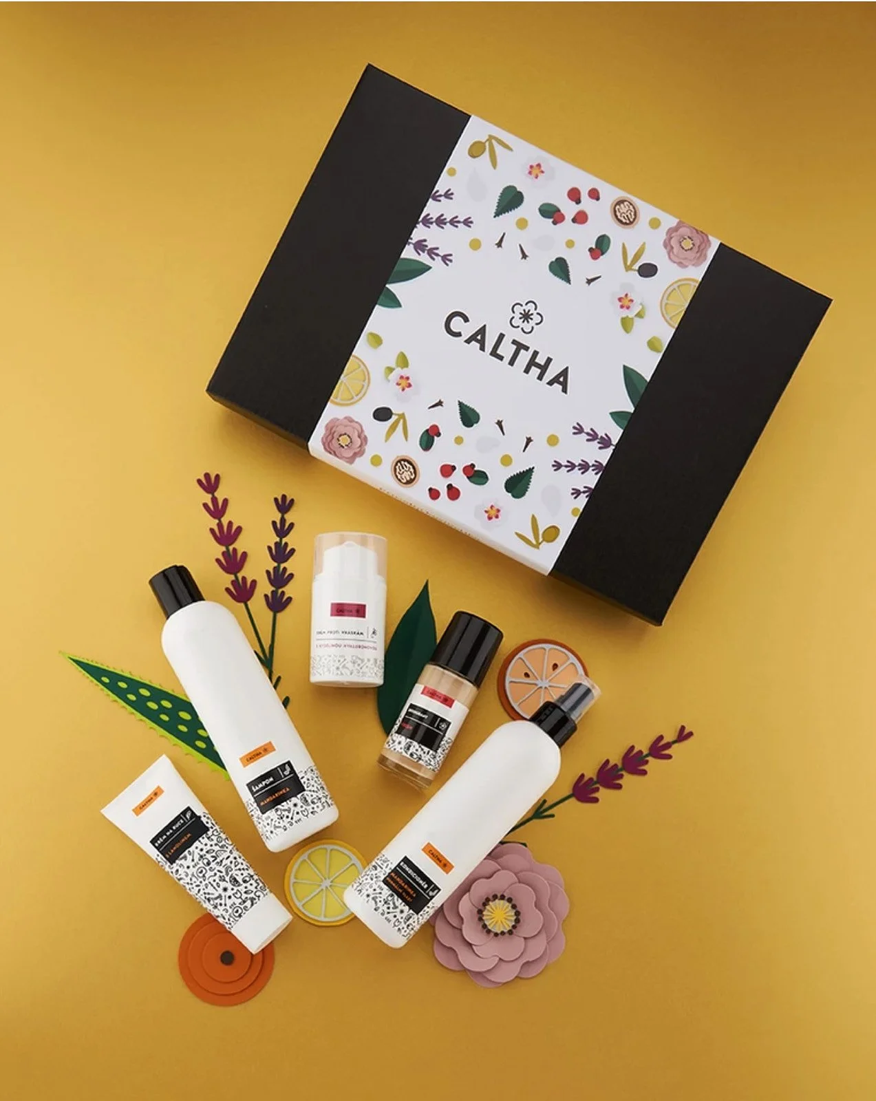 Beauty product set including bottles, tubes, and containers with labels, placed on paper flowers and surrounded by decorative paper cutouts of flowers and lemon slices, with a black and white box with 'CALTHA' in the background.