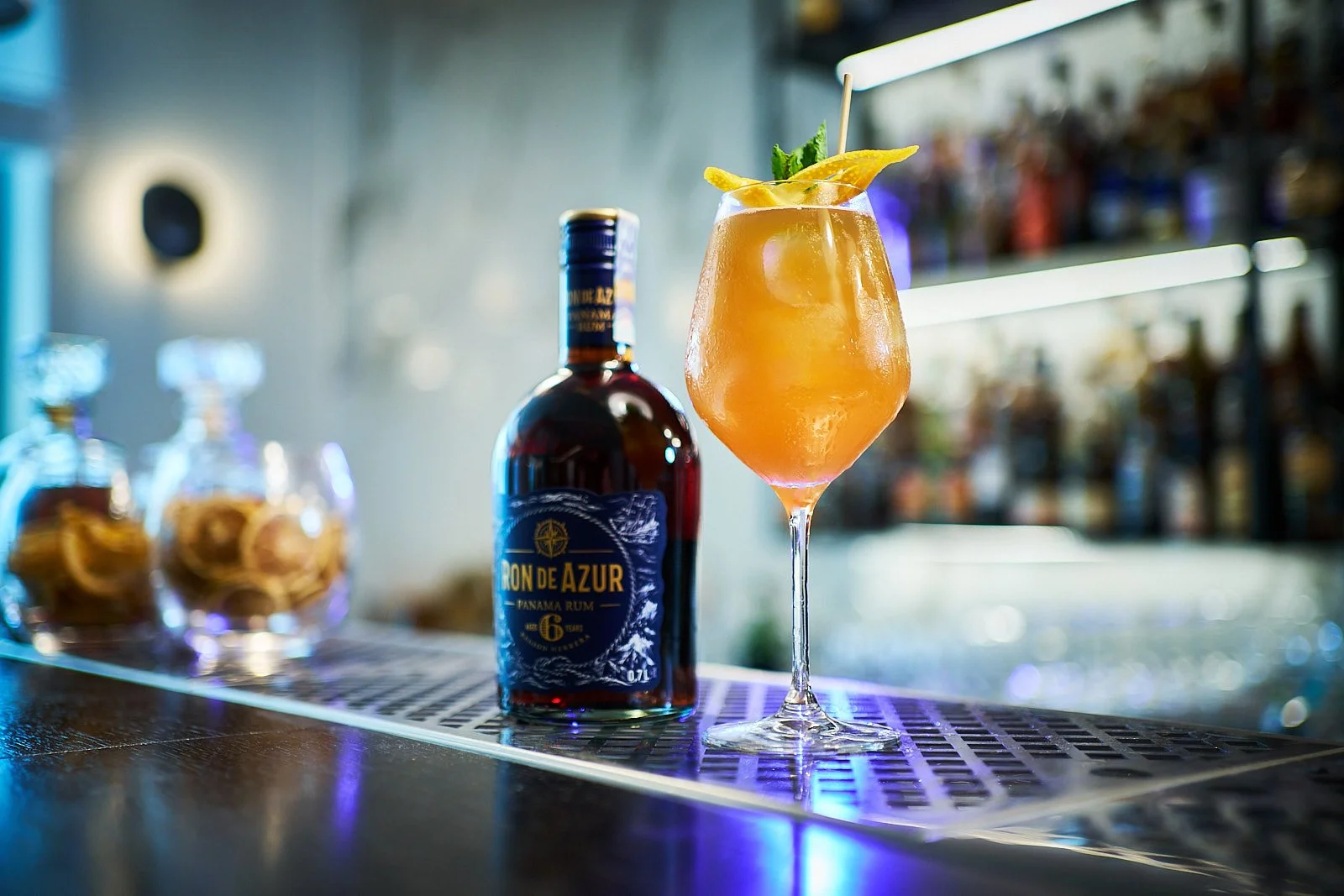 A cocktail in a wine glass garnished with a lemon wedge and mint leaves, with a bottle of Ron de Azur Panama Rum beside it on a bar counter.