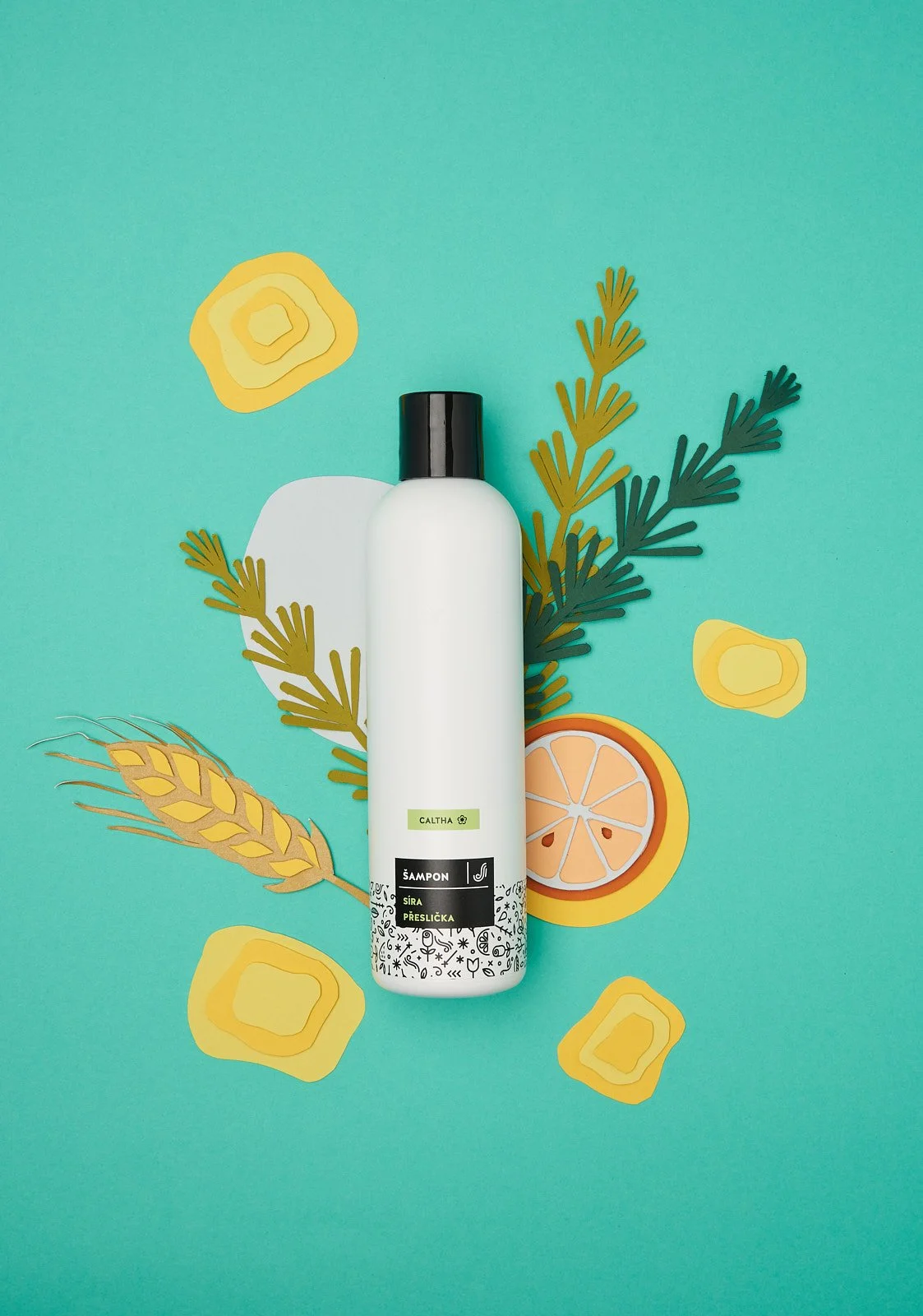 A white shampoo bottle with a black cap on a turquoise background. Surrounding the bottle are illustrated paper cutouts of yellow flowers, green foliage, wheat, and a sliced orange.