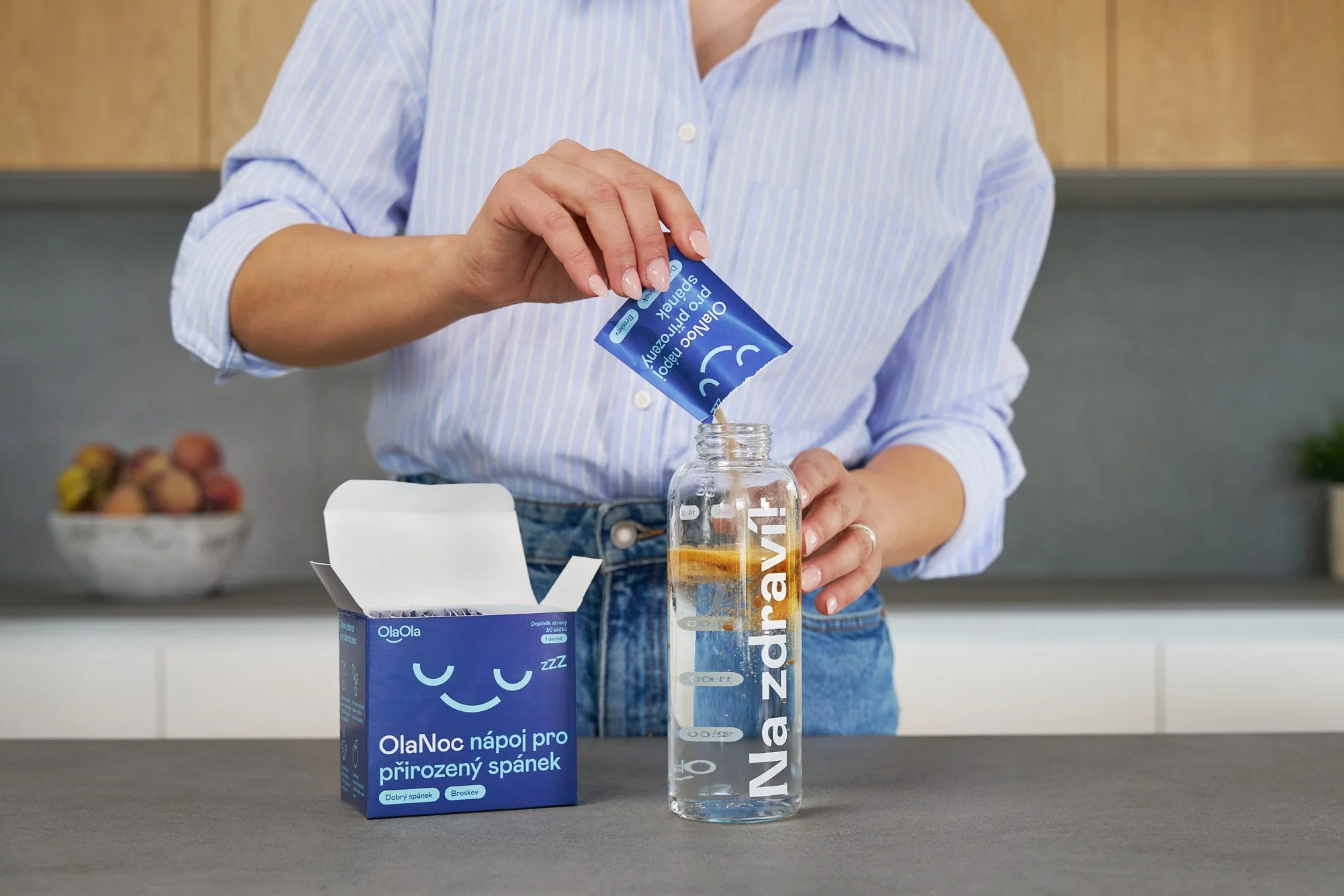 A woman pouring a blue packet labeled 'OlaNoc' sleep aid into a glass water bottle with water. The water bottle has 'Na zadravít' written on it. There is an open box of OlaNoc sleep aid pills on the countertop.