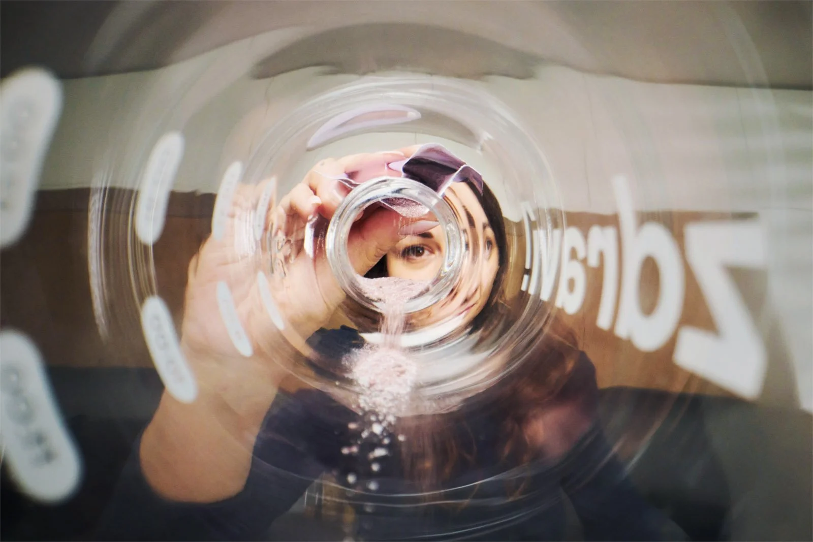 A woman is looking through the opening of a glass container, capturing her reflection and face as she pours pink powder into the container.