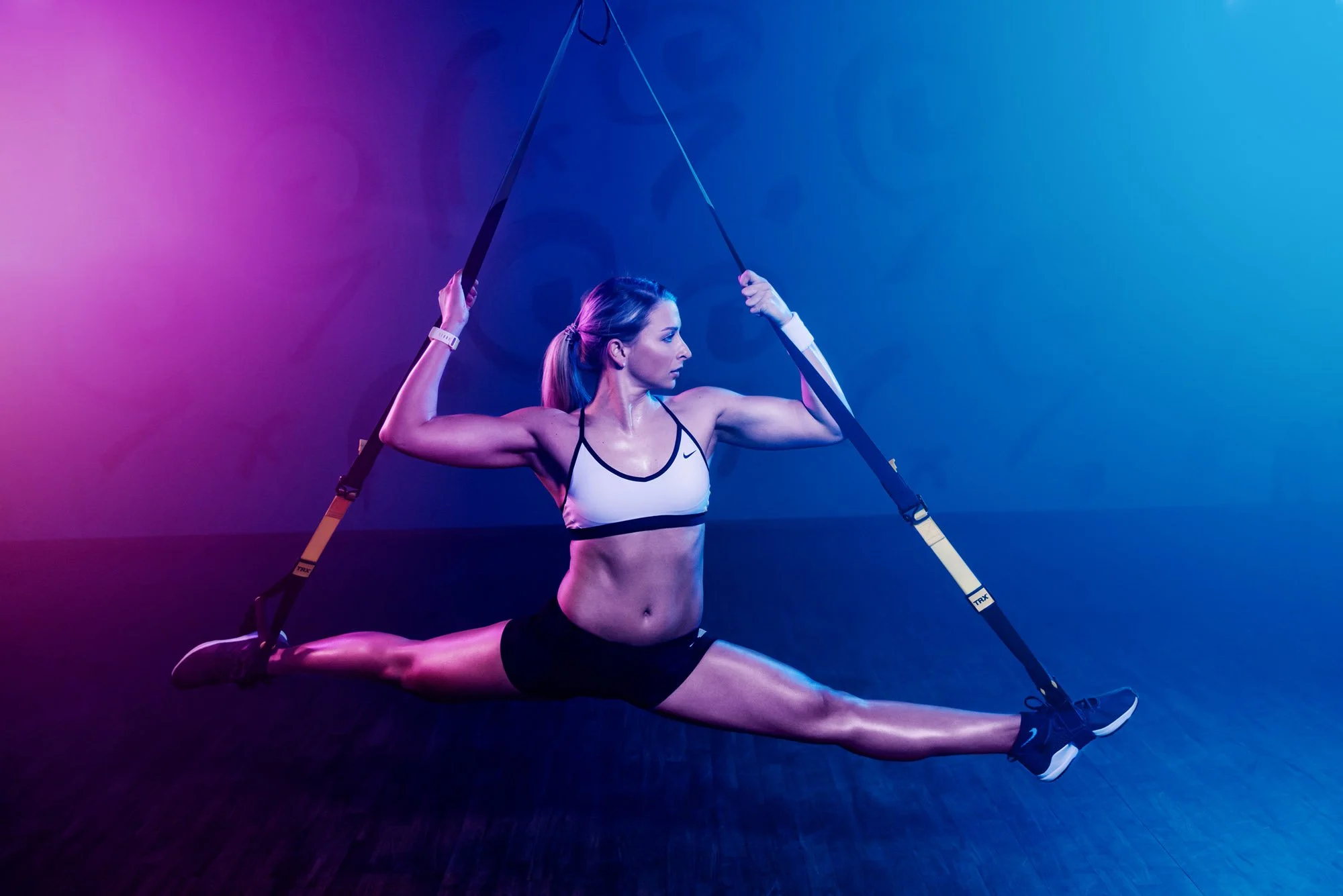 A woman in athletic wear is performing an aerial split with a resistance band in a studio with purple and blue lighting.