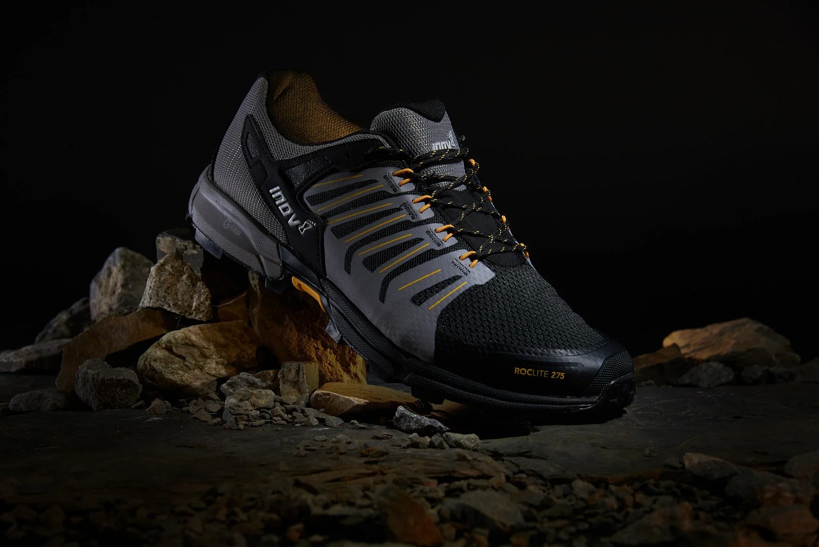 A black and gray trail running shoe with yellow accents, placed on rocks against a dark background.