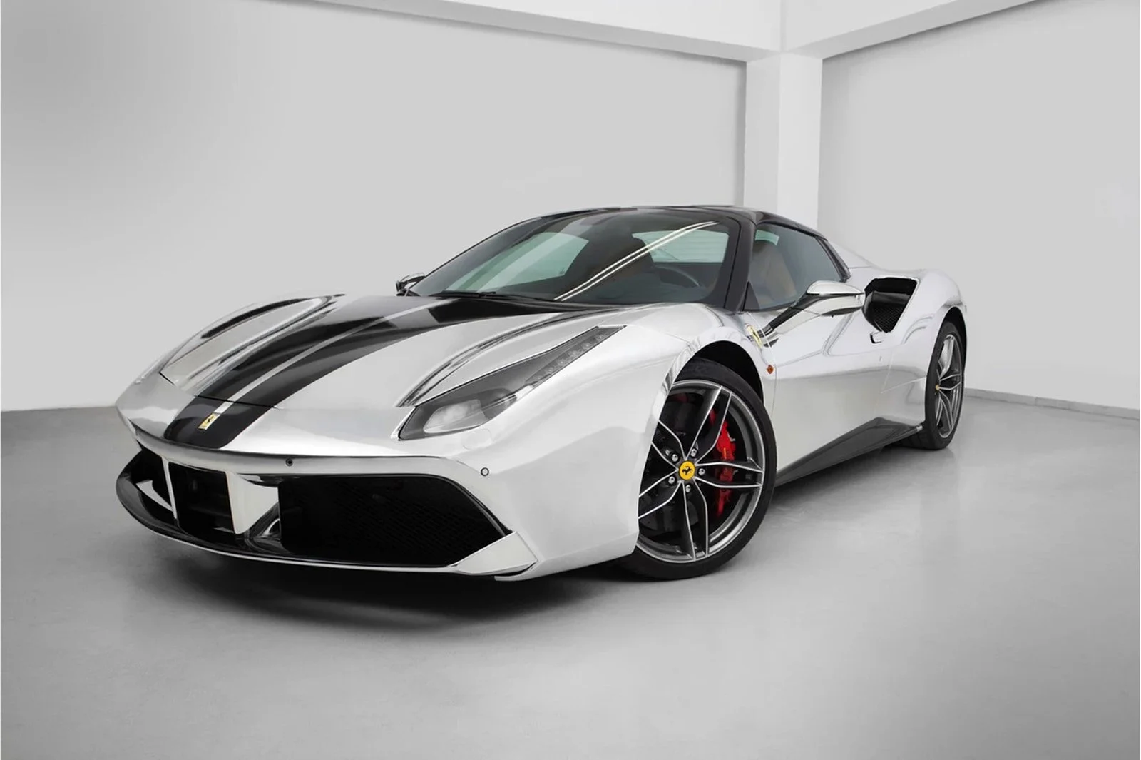 A silver sports car with black racing stripes, low profile, and aerodynamic design, parked indoors with a plain white background.