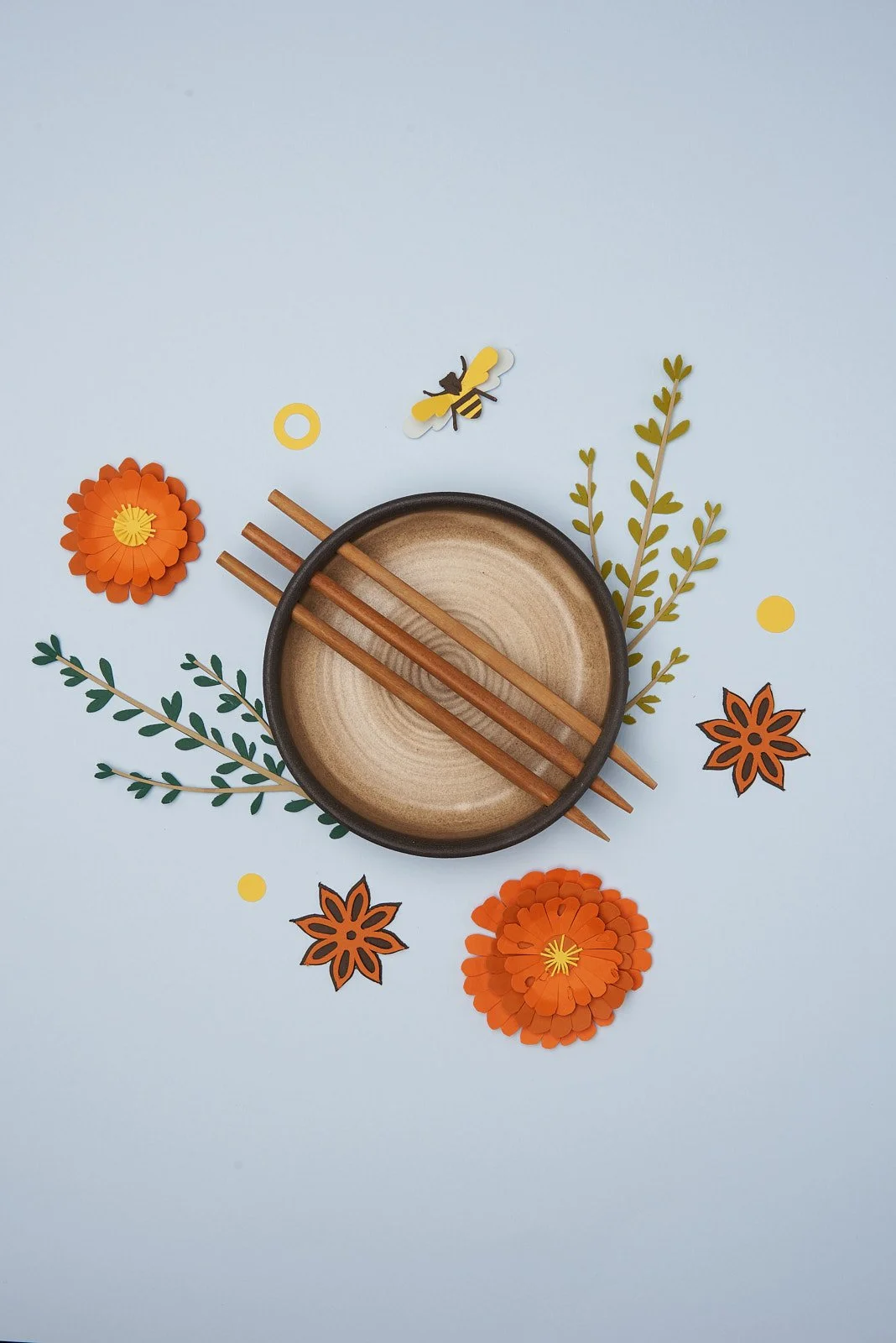 A round wooden bowl with three chopsticks inside, surrounded by paper flowers, green leaves, and paper bee and star decorations on a light blue background.
