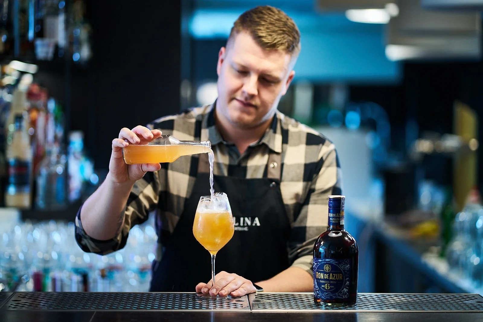 Bartender in a checkered shirt and apron pouring a yellow cocktail from a shaker into a glass with ice, on a bar counter with a bottle of Ron de Azur rum nearby.