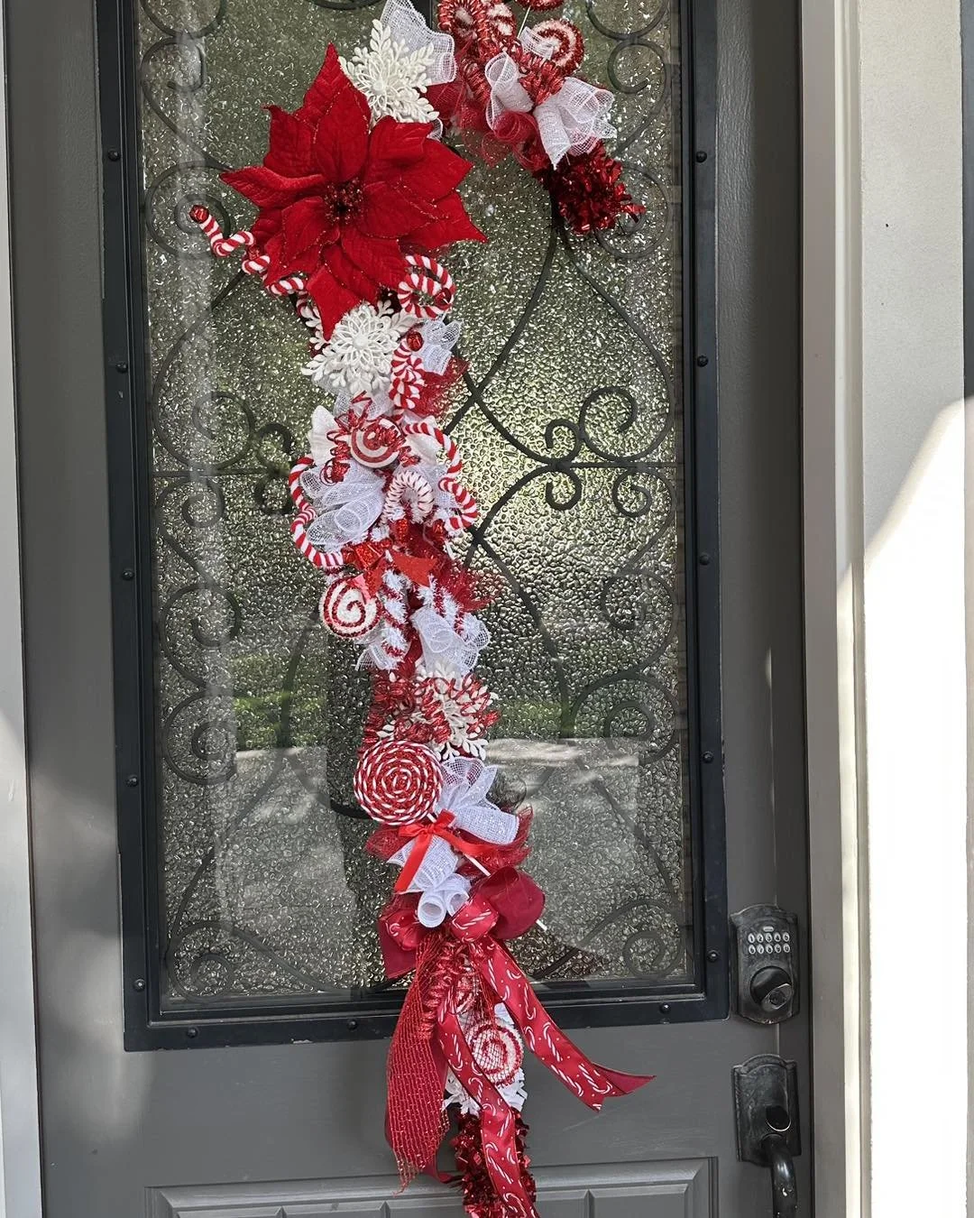 Decorative holiday wreath with red poinsettia flower, white and red ribbons, candy cane patterns, and ornaments hanging on a front door with textured glass.