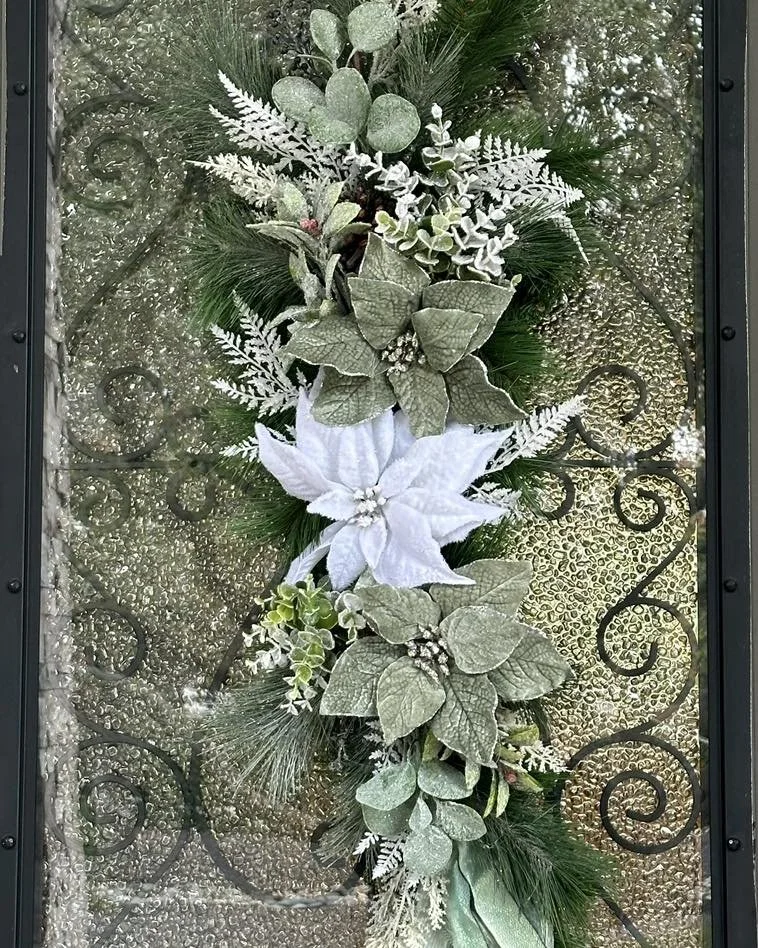 Decorative holiday wreath with green pine branches, sliced leaves, white poinsettia flower, and other frosted foliage against a textured glass background.