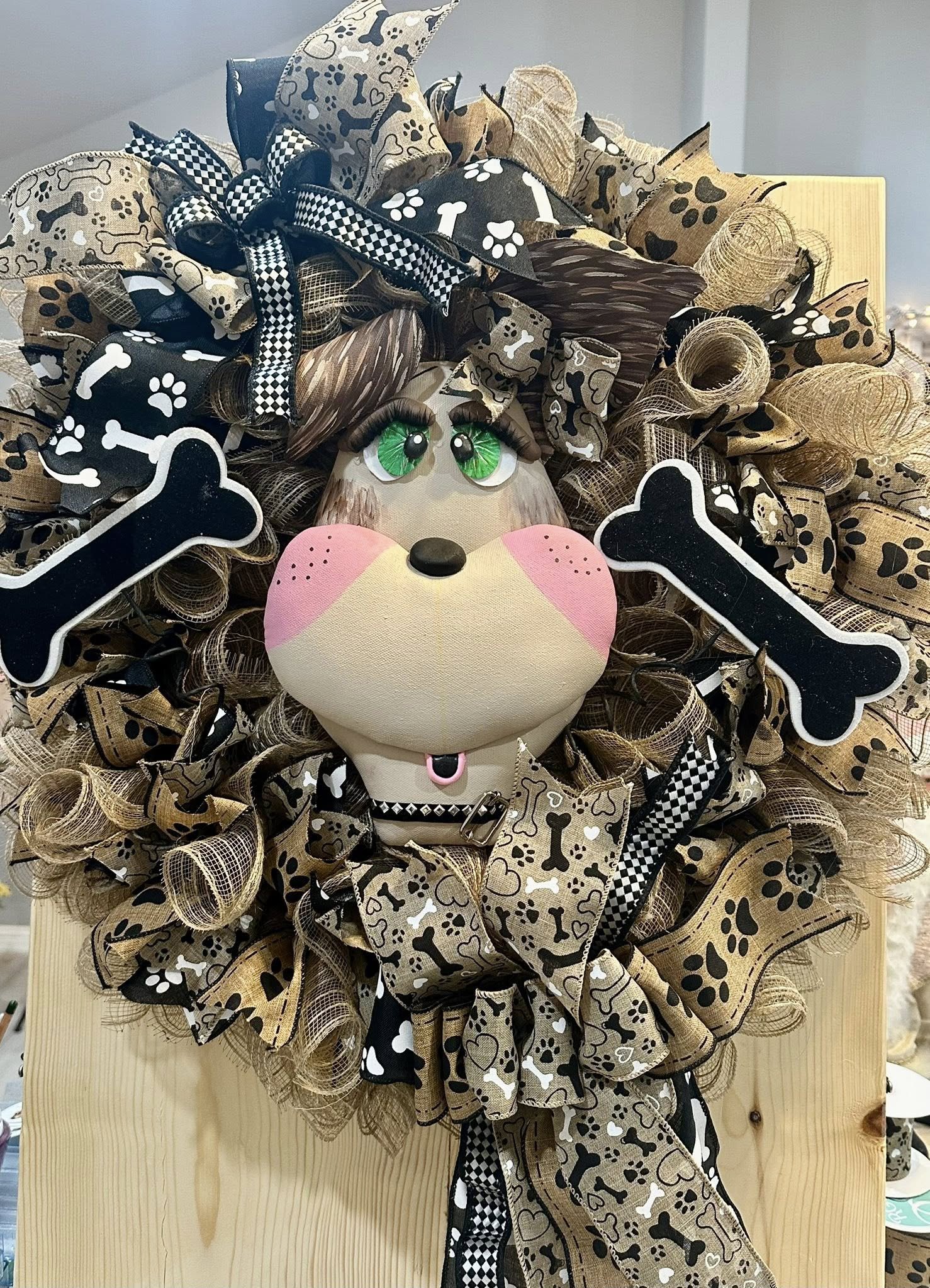 Decorative wreath shaped like a dog with a face, big green eyes, pink cheeks, and a black nose, made of ribbons with bone and paw print patterns, and adorned with black bone-shaped cutouts.