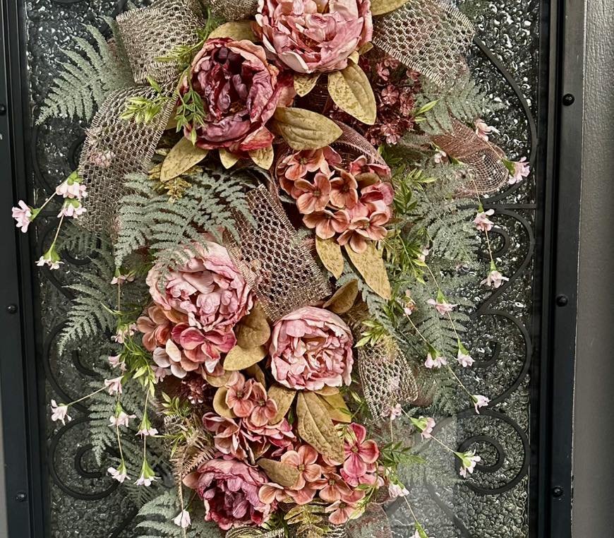 Decorative floral arrangement with pink peonies, pink and peach hydrangeas, green leaves, fern fronds, small pink flowers, and a mesh ribbon, displayed on a decorative iron gate.