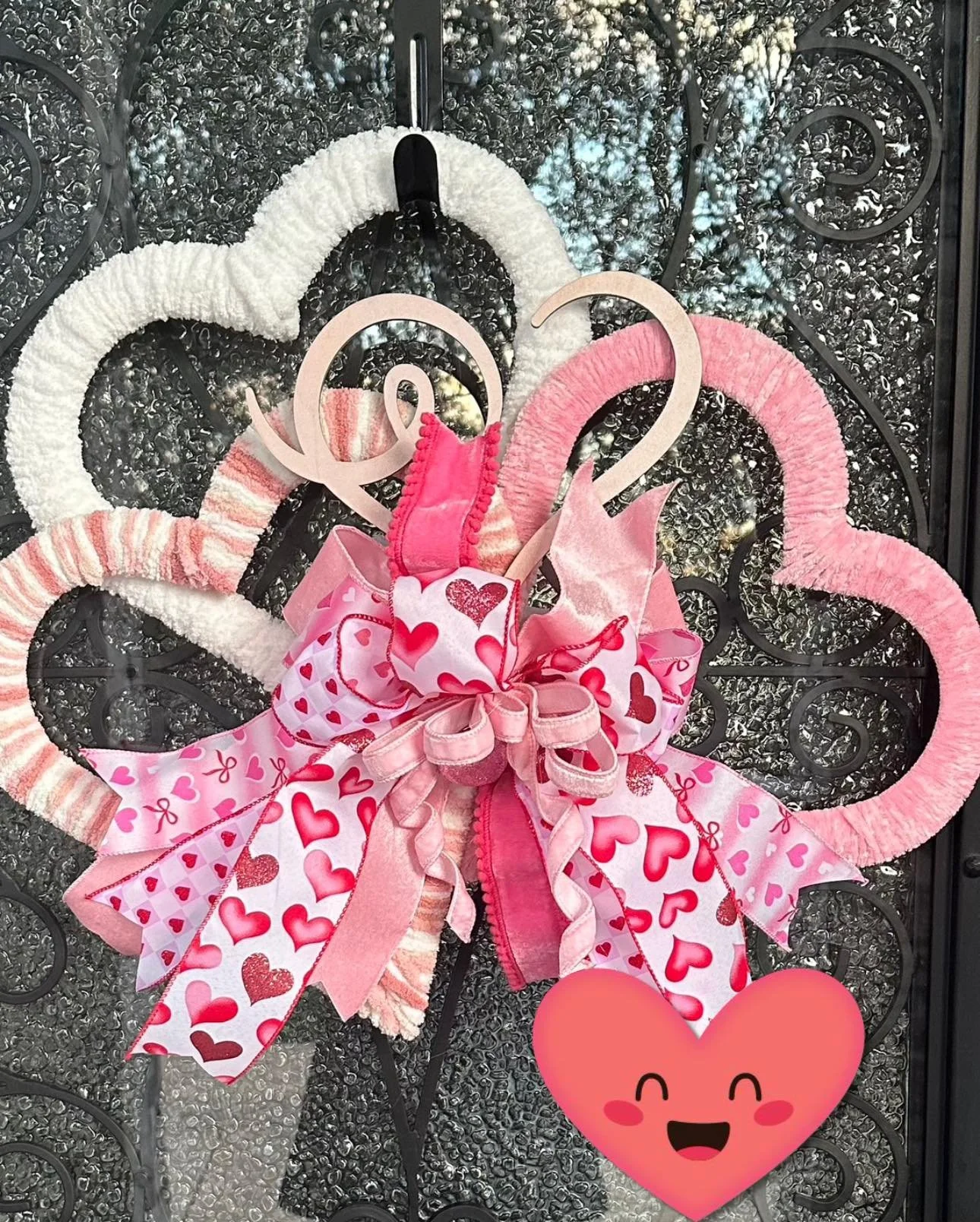 Decorative heart-shaped wreath with pink and white fabric, ribbons, and heart patterns, mounted on a black wrought iron gate with a cartoon heart emoji at the bottom.