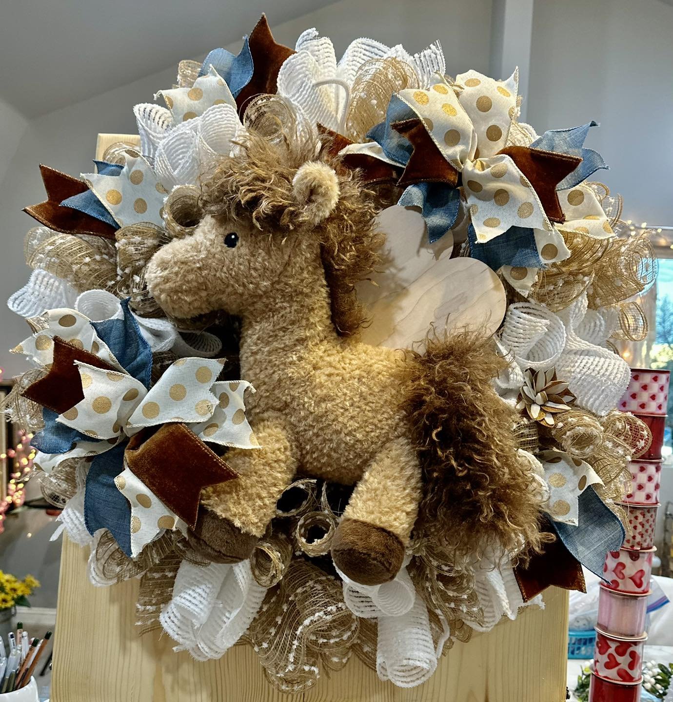 A decorative plush horse surrounded by various ribbons and mesh fabric in gold, white, blue, and brown, arranged in a circular fashion on a wooden display.