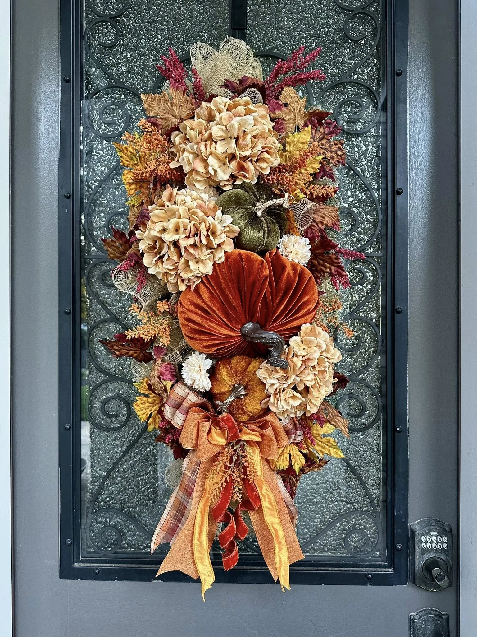 Fall autumn-themed door wreath with artificial pumpkins, hydrangeas, fall leaves, ribbons, and decorative accents on a glass pane door.