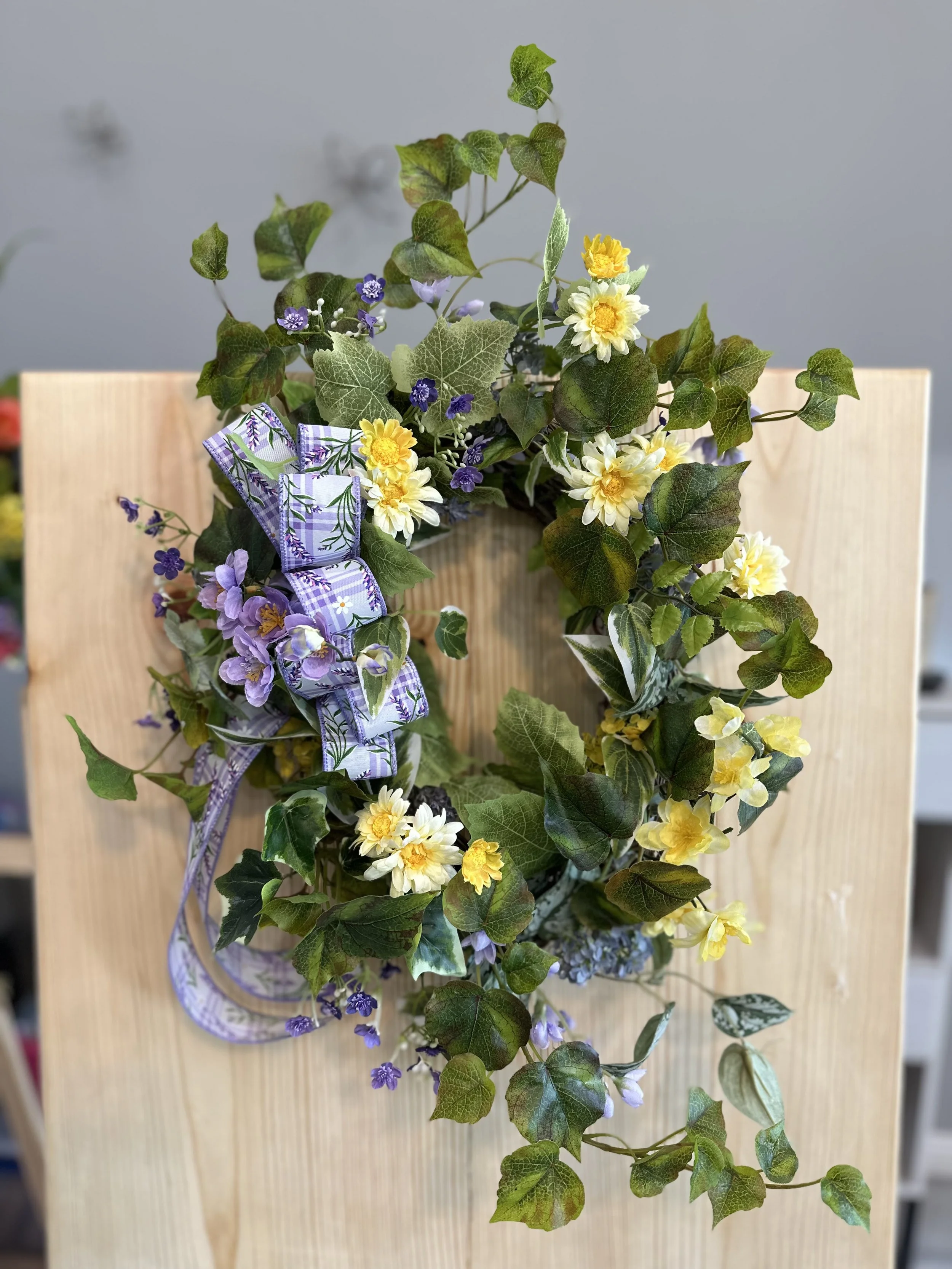 A decorative floral wreath made of ivy leaves, yellow and white flowers, purple flowers, and a purple ribbon with a floral pattern, hanging on a wooden surface.
