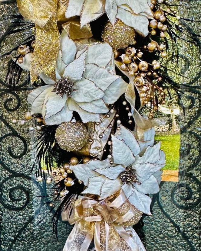 Close-up of a festive holiday wreath decorated with white poinsettias, gold and white ribbons, gold berries, and golden glitter accents. The wreath is hanging on a black wrought iron gate.