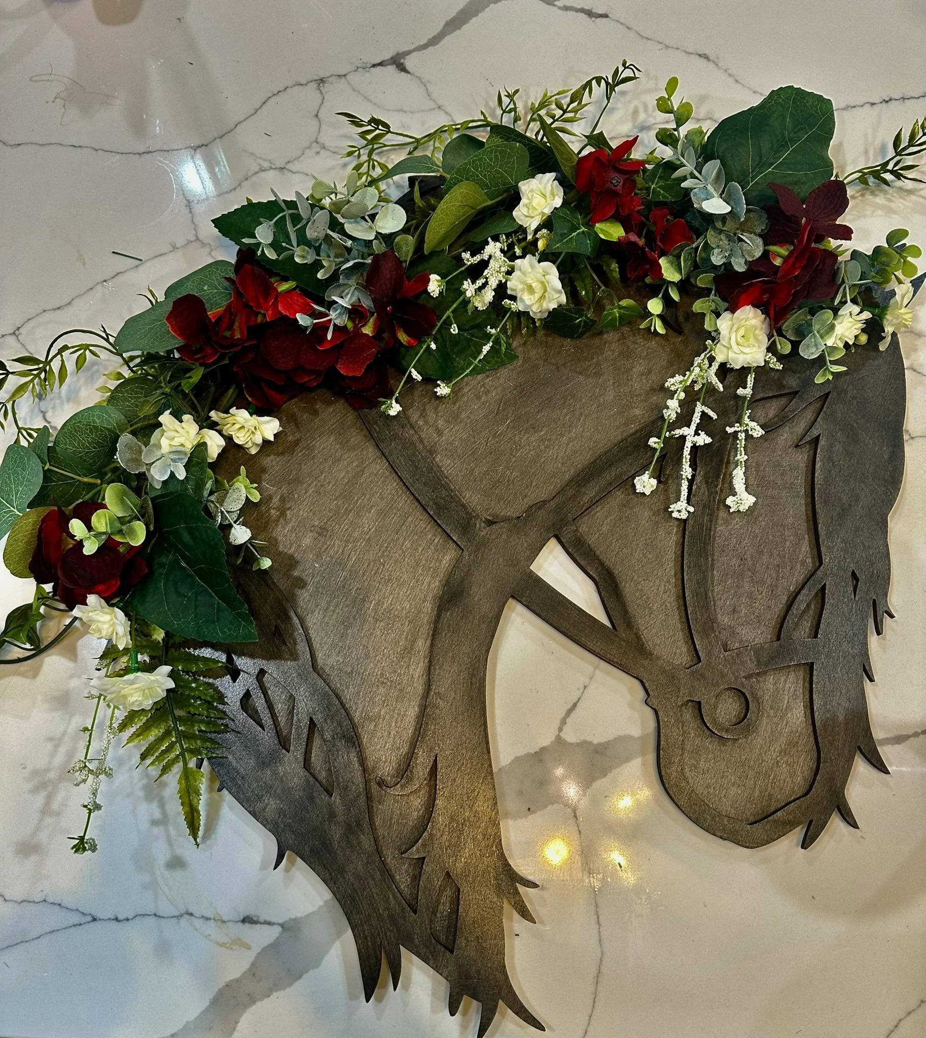 Decorative horse-shaped wall hanging with a floral arrangement of red, white, and green artificial flowers and foliage, mounted on a marble wall.