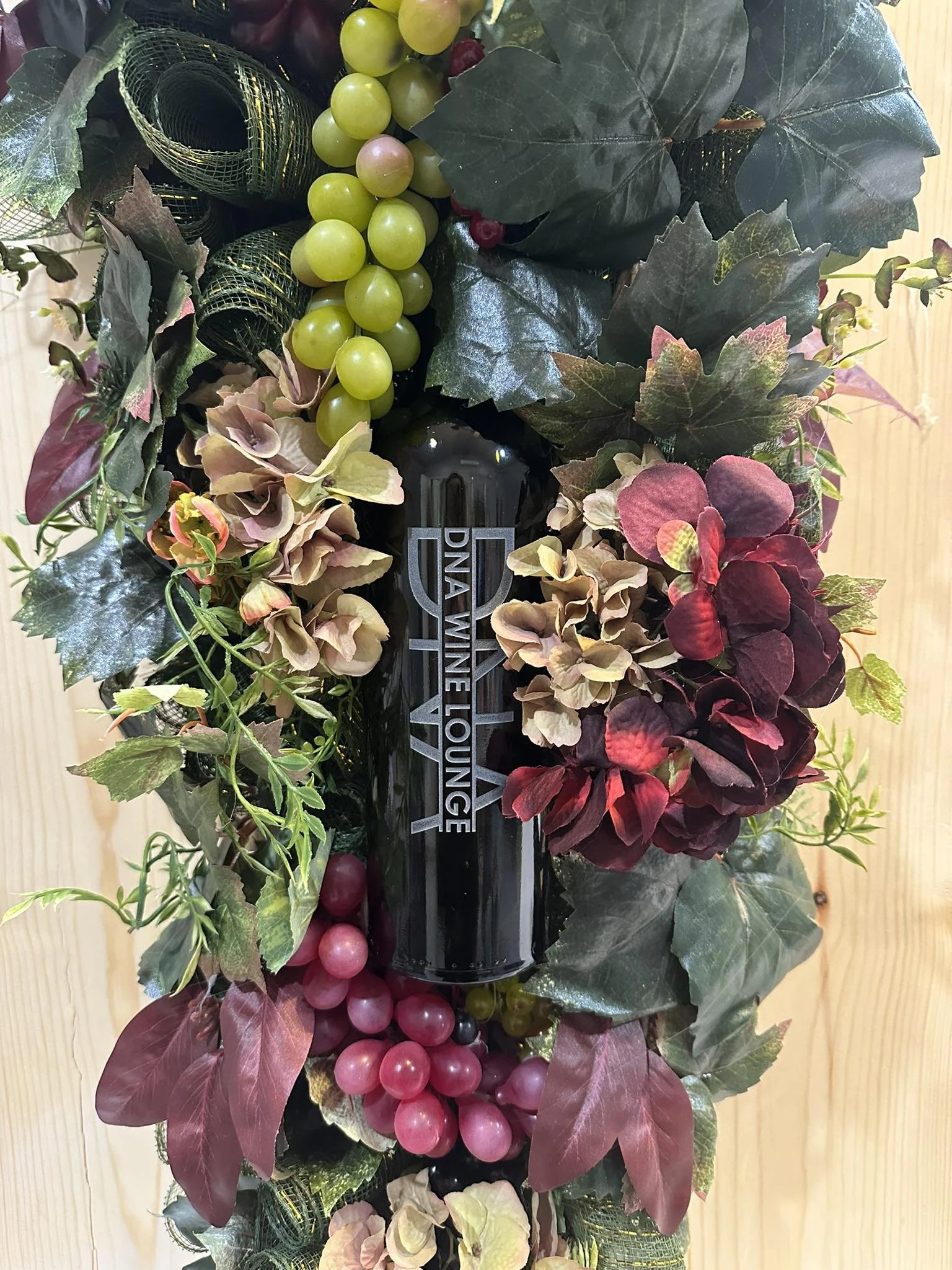 A decorative wreath made of variegated leaves, grapes, and hydrangeas surrounds a black bottle with the words "Diva Wine Lounge" on it, against a wooden background.