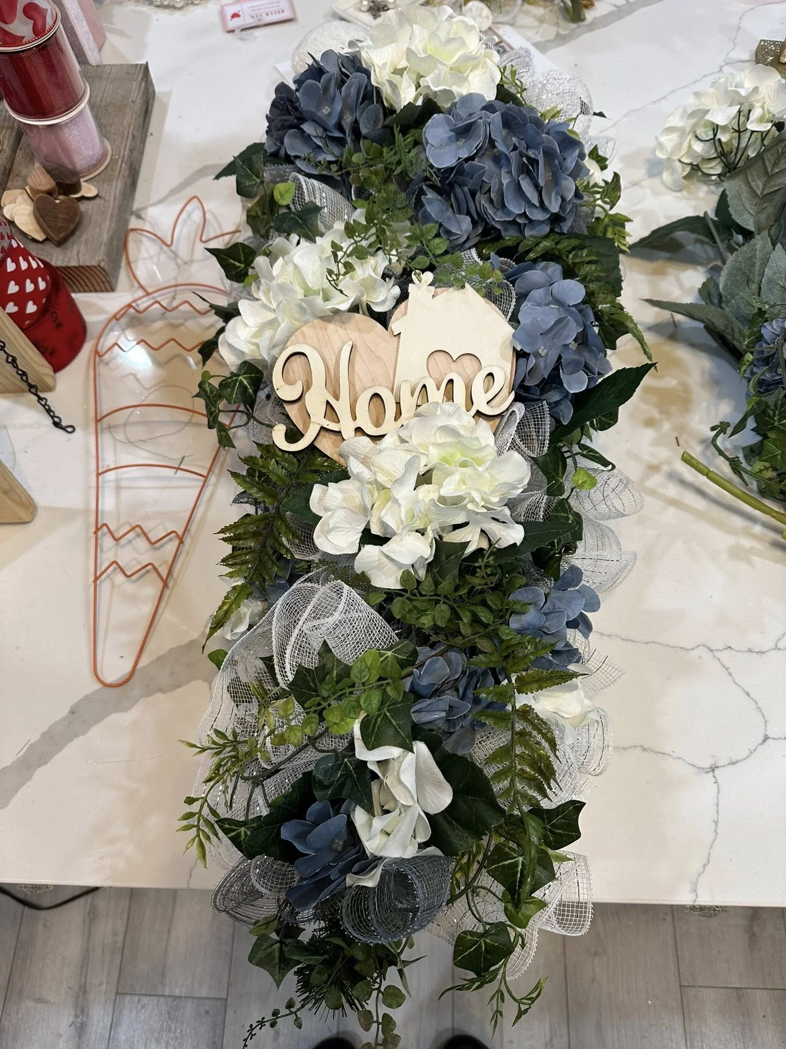 A floral arrangement with white and blue hydrangeas, green leaves, and white mesh ribbons, featuring a wooden sign that says 'Home.'