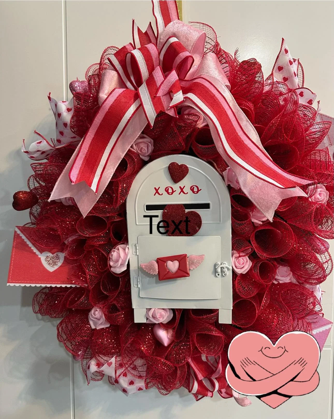 Valentine's Day-themed decorative wreath with red and pink ribbons, heart accents, and a small mailbox decoration with a pink heart envelope. An illustration of two hugging hearts is on the bottom right corner of the image.
