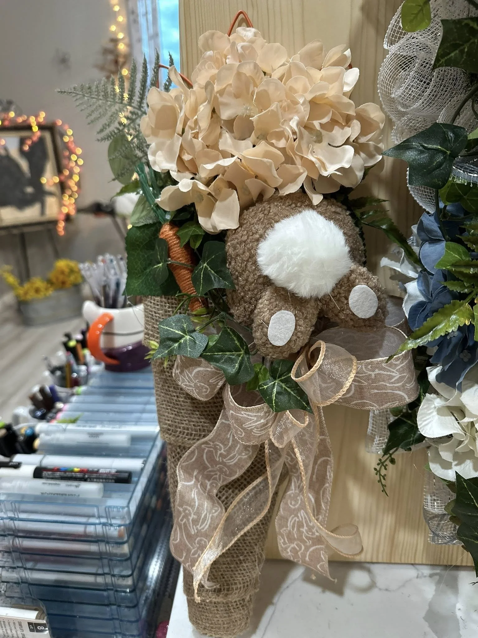 A decorative bouquet featuring beige flowers, green ivy, and a plush teddy bear with a white nose, attached to a burlap-wrapped handle with a sheer ribbon tied into a bow.